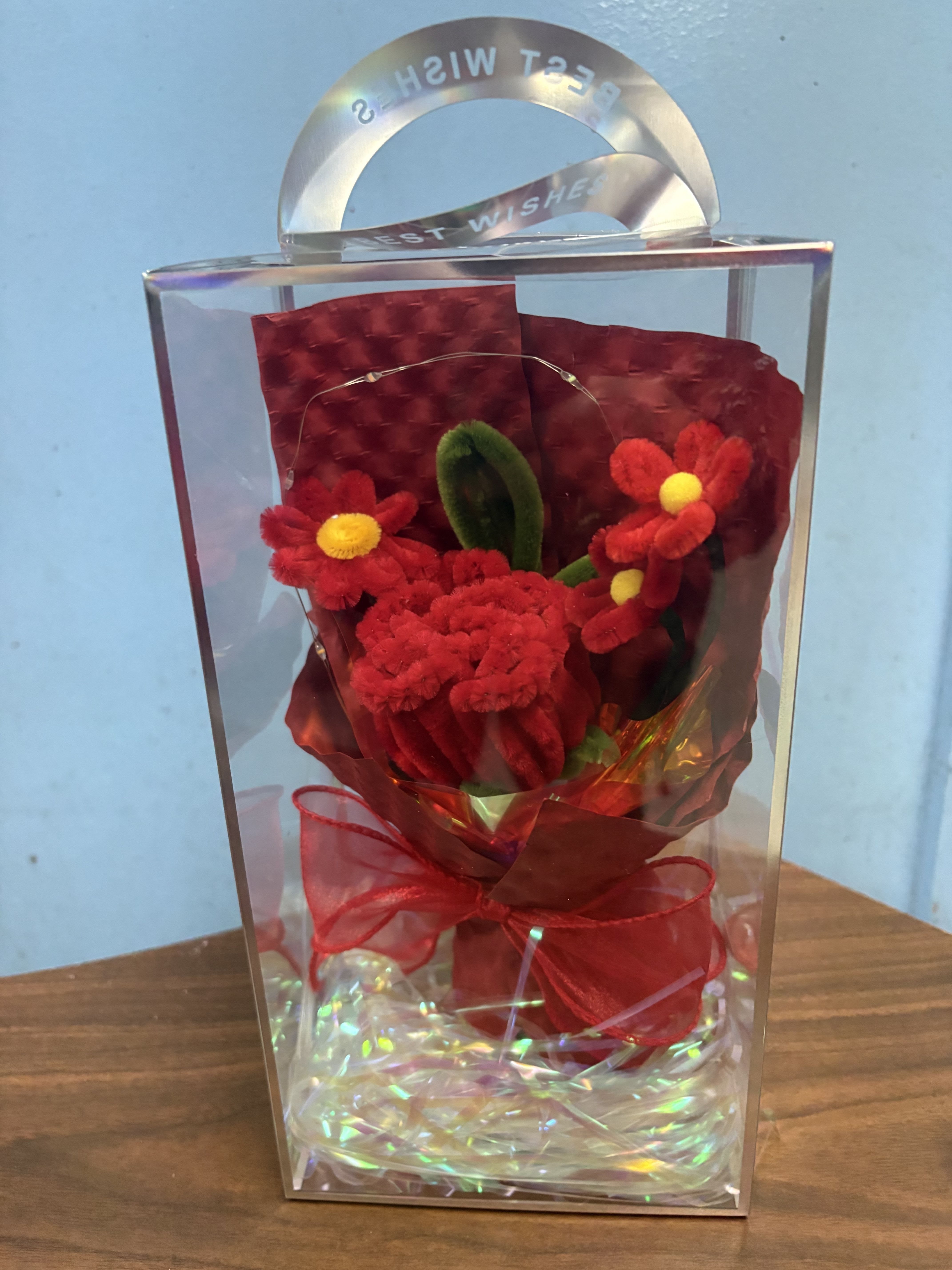 Keepsake Bouquet  - Bright red carnations packed into a clear rectangular gift box make a joyful, romantic statement for anniversaries, Valentine's Day, or just because. This small boxed floral gift reads like a little love letter - the tight red blooms, a few daisy-like red and yellow accents, and a single green leaf pick create a cheerful, affectionate look that will make the person you're thinking of feel truly seen.  Presented in a transparent plastic keepsake box with iridescent filler, decorative cellophane wrap, and a red ribbon bow, the arrangement arrives ready to gift and display. A metal handle makes it easy to carry, and the compact size is perfect for birthdays, bedside tables, or a desk to brighten someone's day. Designed to convey romance and warm celebration in an elegant, no-fuss package, it's an effortless way to celebrate love, a milestone, or simply make someone smile.