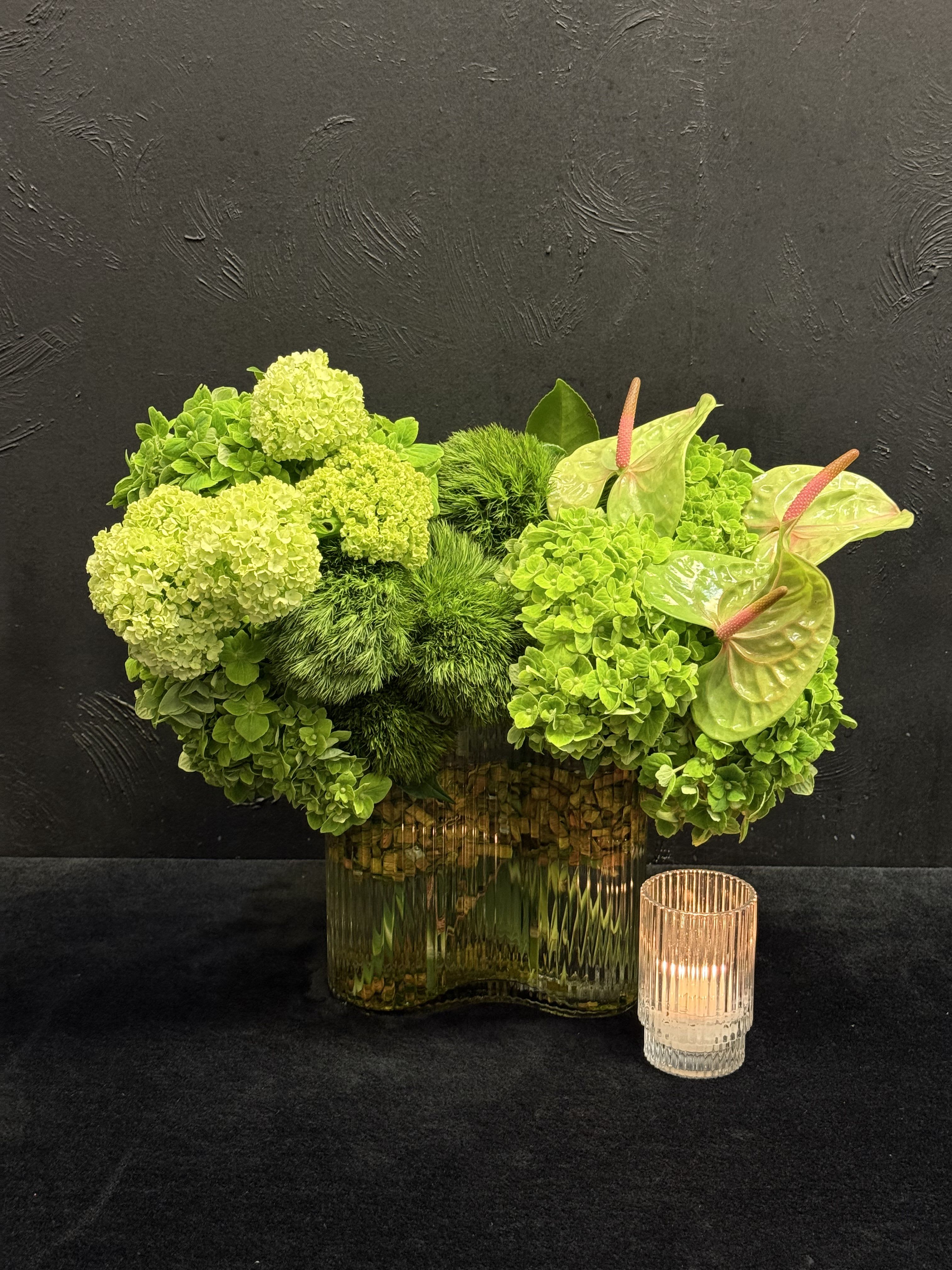 Green floral arrangement in a glass vase with an anthurium and candle