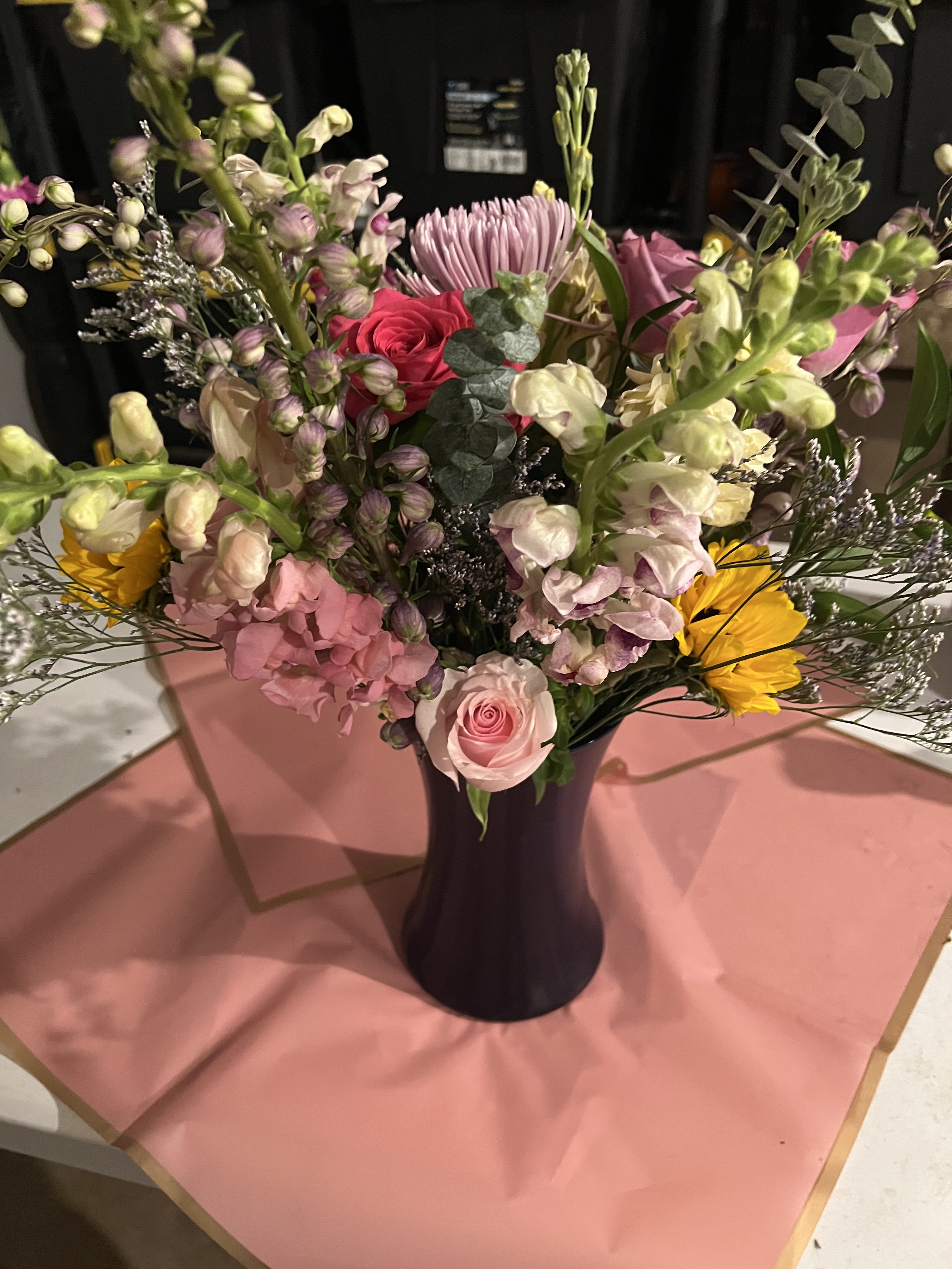 Spring Melody - A lively, garden-style flower arrangement that sings of spring-perfect as a birthday bouquet, a cheerful thank-you gift, or an everyday surprise to brighten someone's day. Spring Melody pairs soft pink roses and snapdragons with creamy hydrangea clusters, lavender chrysanthemums, sunny yellow daisy-like blooms and a pop of bright pink carnation so the recipient feels genuinely celebrated the moment they see it.  The arrangement is designed with contrasting forms-rounded roses and hydrangeas against the vertical snapdragons and airy filler-to create both fullness and movement. Eucalyptus and light filler stems add height and soft texture, and the design is offered in a choice of vases (dark ceramic, cobalt blue glass, or a lighter ceramic) to suit the recipient's style. Sized to sit beautifully on a table or mantel, it's an inviting, versatile choice for spring occasions and everyday gifting; simple care-fresh water and a cool spot-keeps the blooms looking their best.