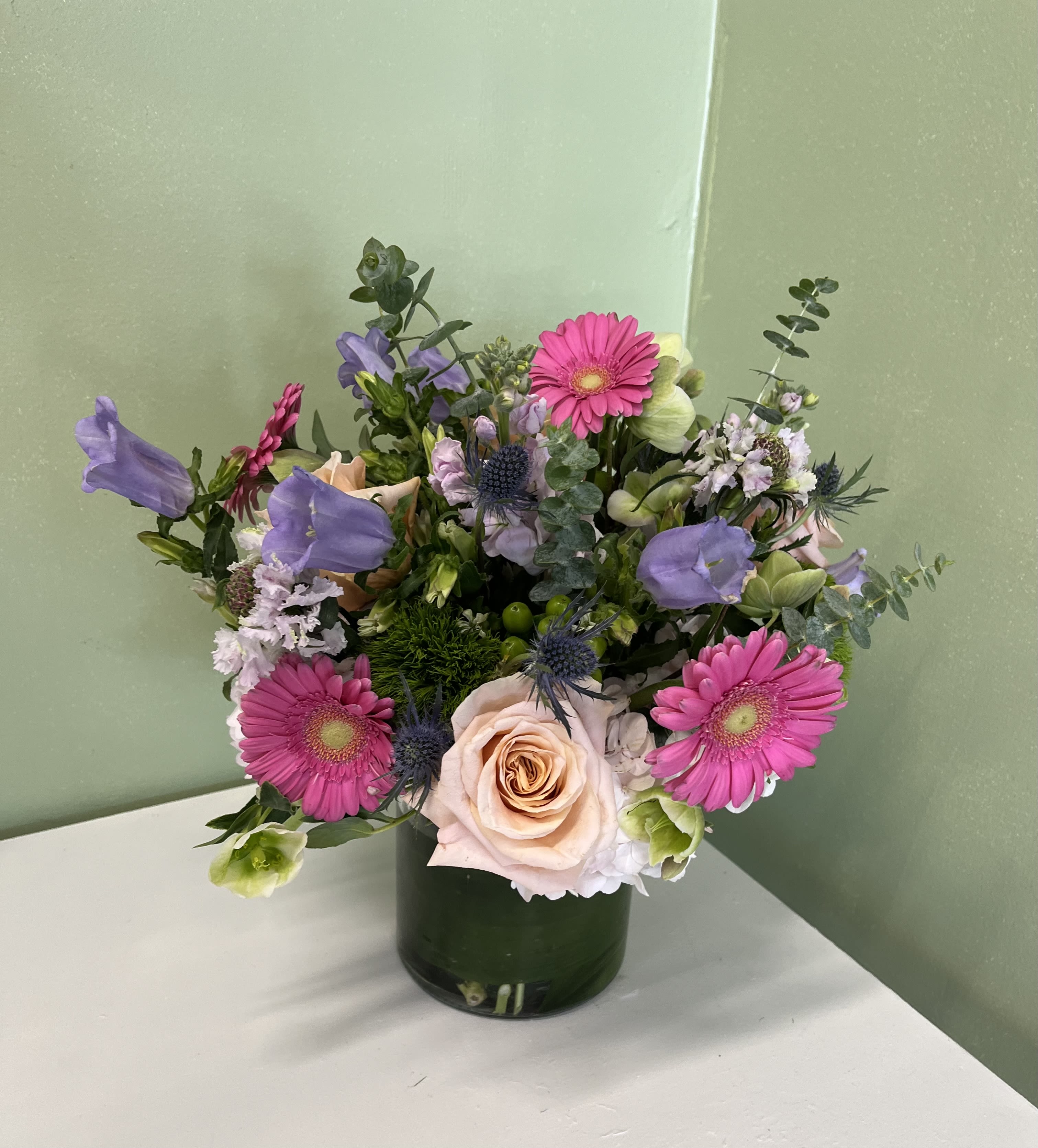 Mixed arrangement of pink gerbera daisies, peach roses, and purple blooms in a green glass cylinder vase