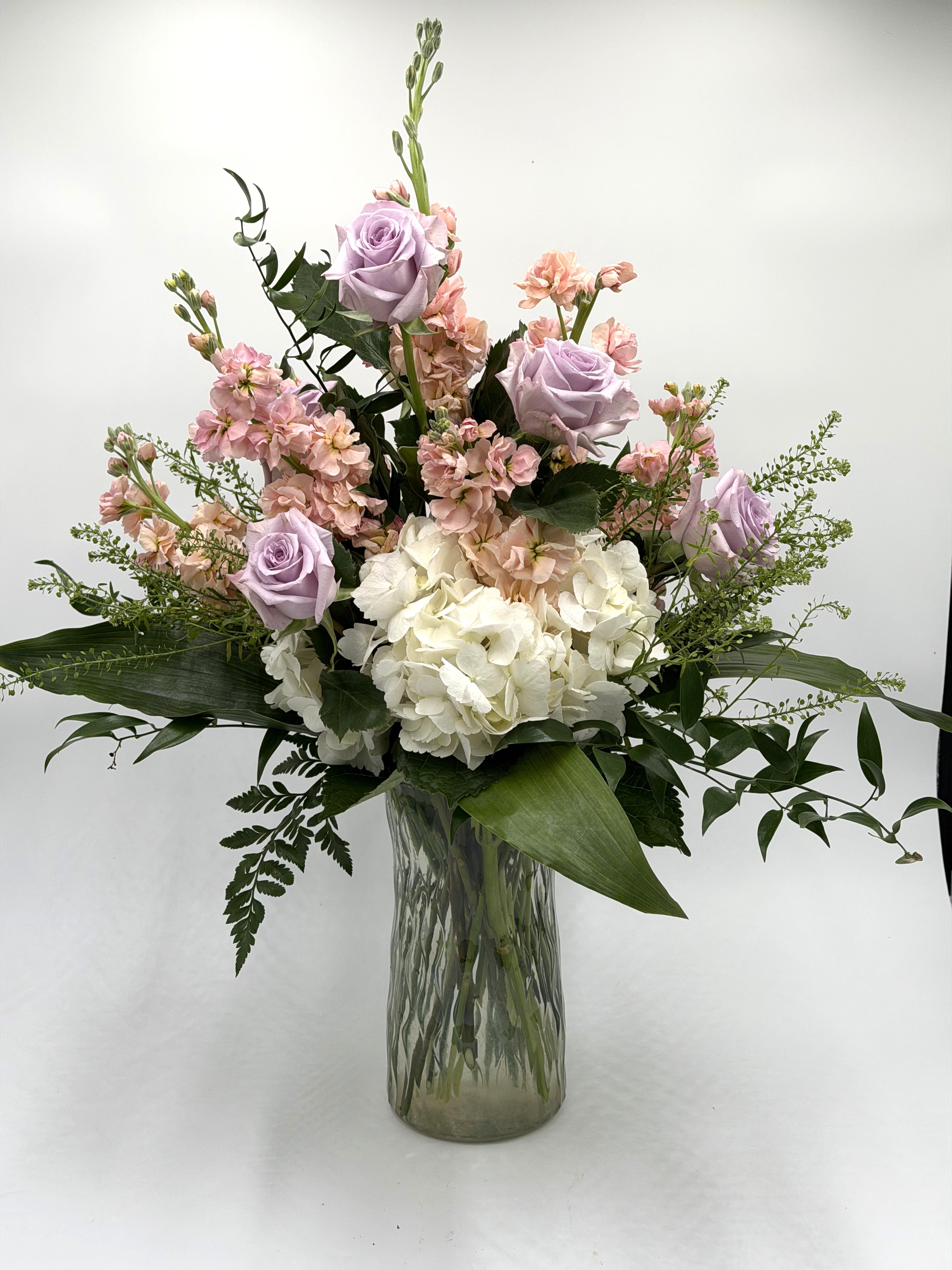 Softly Spoken Beauty - This elegant arrangement features soft  roses, blush stock, and lush white hydrangea for a refined, garden-inspired look. Airy greenery and natural movement give the design a fresh, organic feel with beautiful height and dimension. Arranged in a textured glass vase, it’s a timeless statement piece perfect for celebrating  with sophistication and love.