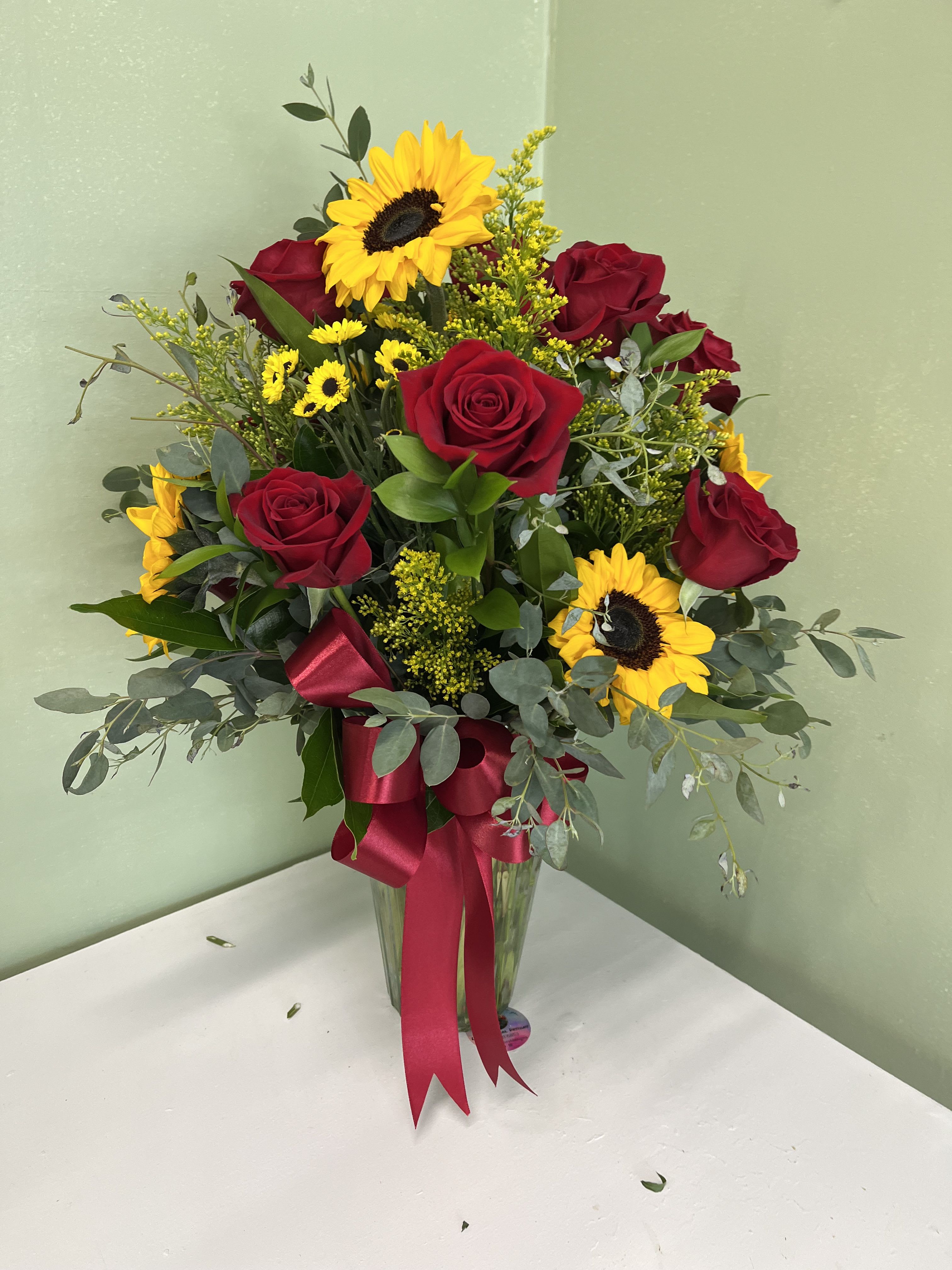 Tall vase of red roses and yellow sunflowers with small yellow daisies and a red bow.