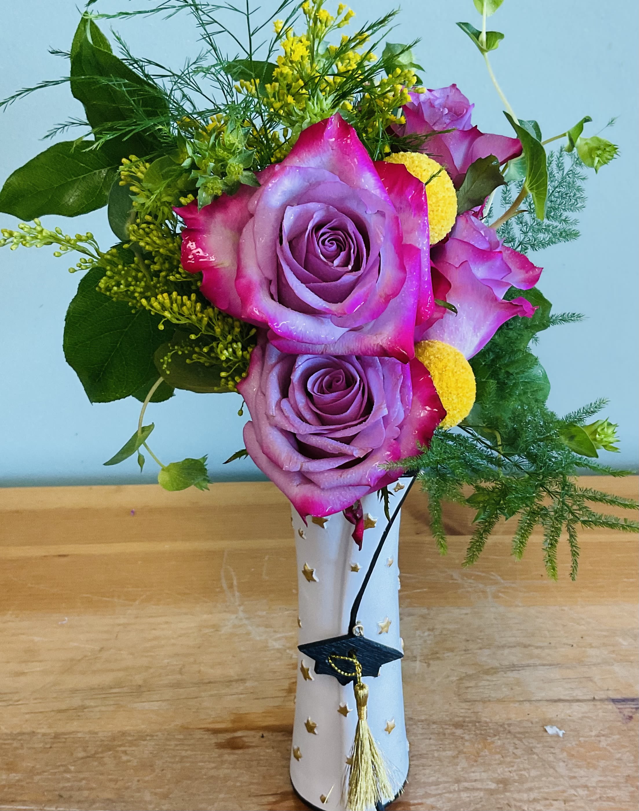 Brainy Blooms - Vivid purple roses edged in bright pink make a joyful statement in this graduation bouquet, the perfect congratulatory arrangement for a graduate. Designed for graduation or celebration, the cheerful color contrast and festive accents read as both playful and proud - the kind of gift that makes the recipient feel seen and celebrated on their big day.  The bouquet mixes large purple-pink roses with small yellow button blooms and airy green filler for a textured, upbeat look. Flowers arrive arranged in a white vase decorated with gold stars, and a removable graduation cap charm with a gold tassel hangs at the front for an unmistakable graduation touch. The compact vase keeps the display neat on a dorm shelf or mantel while letting the vivid colors take center stage.  This arrangement suits recent grads, proud parents, or anyone marking academic milestones - a meaningful way to say "congratulations" that looks great in photos. Vase dimensions and care: keep water fresh, trim stems every few days, and place away from direct heat to extend bloom life.