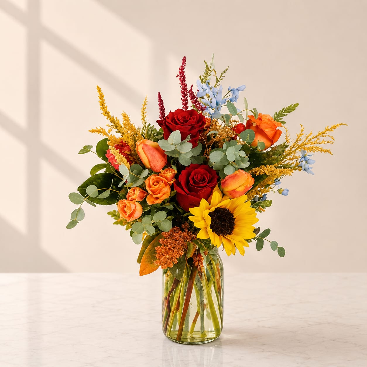 Because of You - Seasonal Fall Bouquet  - Because of You – A Vibrant Floral Gift of Gratitude  Brighten someone’s day and show just how much they mean to you with Because of You, a joyful bouquet bursting with color and life. This stunning flower arrangement features radiant orange and red roses, cheerful orange tulips, striking blue delphinium, and fresh eucalyptus—all beautifully arranged in a charming clear mason jar. Perfect for expressing gratitude, celebrating special moments, or simply surprising a loved one, this modern and uplifting design is guaranteed to bring smiles. Send Because of You today and let your heartfelt appreciation bloom in the most memorable way.