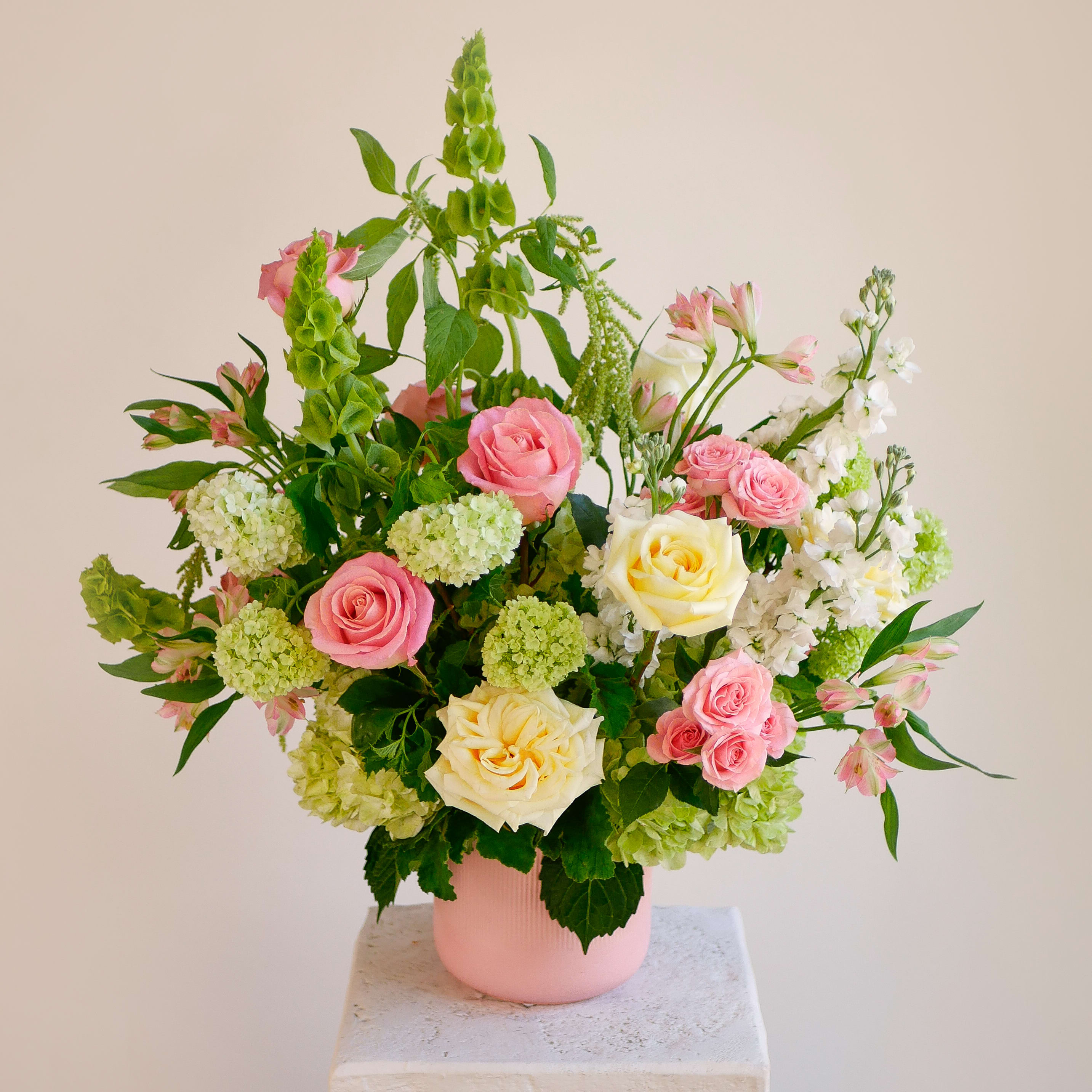 Sweetest Devotion - Pink and cream garden roses are the focal point of this garden-style arrangement, accented by pink spray roses, white stock, and delicate alstroemeria. Green vibrunum, hydrangea and architectural bells of Ireland provide structure and a fresh, verdant base. Arranged in a matte pink ceramic vase, the piece measures approximately 28 inches tall and 25 inches wide, offering an elegant grand presence. Every stem is hand-selected for color harmony and longevity. Local same-day delivery is available across West LA.