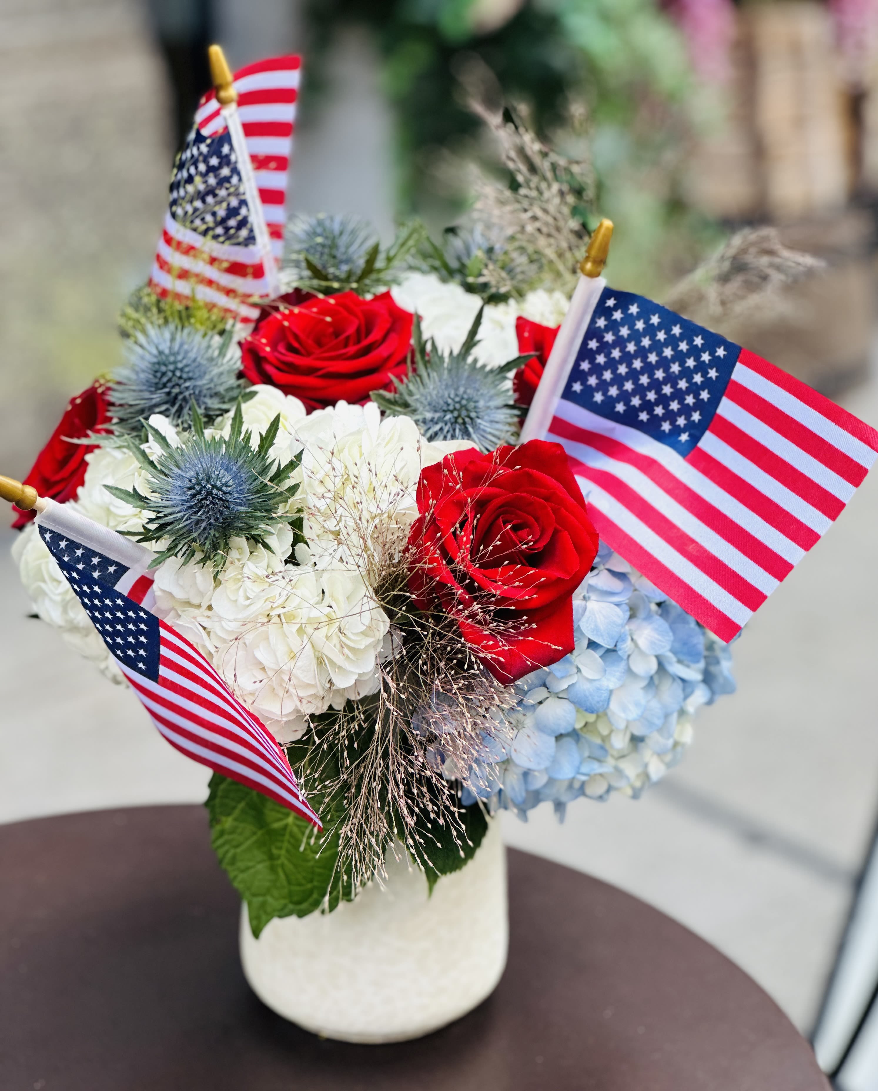 Red roses and blue hydrangeas in a white vase with small American flags