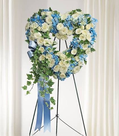 Blue Eternity Heart - An elegant open-heart standing tribute designed in serene blue and crisp white blooms, accented with a flowing blue ribbon to symbolize everlasting love, peace, and remembrance. This heartfelt arrangement features a sophisticated blend of white roses, chrysanthemums, blue hydrangeas, and white carnations, beautifully framed with fresh greenery in an open-heart design. Displayed on a standing easel, the tribute offers a meaningful expression of sympathy, honoring a life with grace, loyalty, and deep admiration