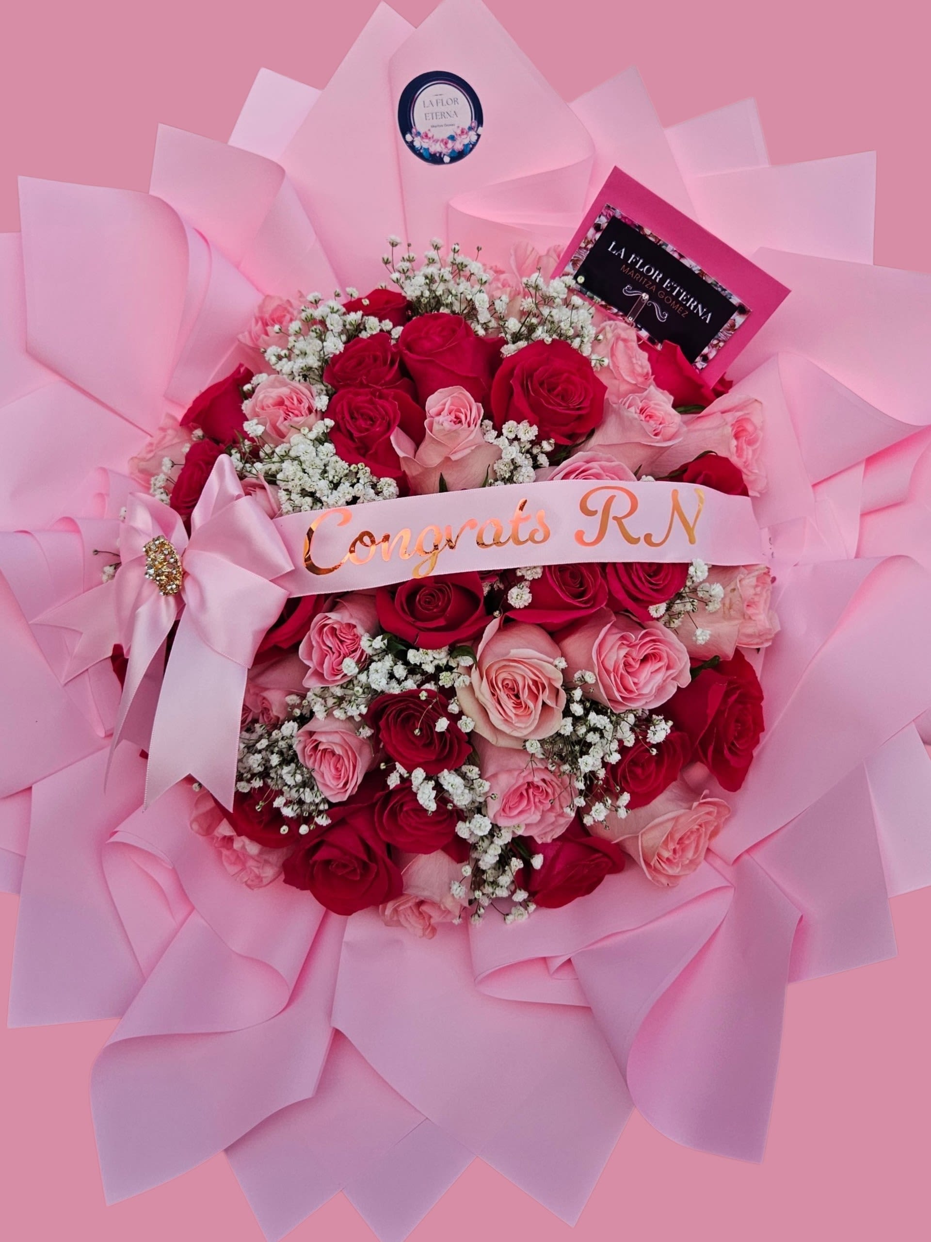 Congrats  RN  - A lush cascade of red and blush-pink roses says celebration the moment she sees it-this congratulatory flower arrangement is perfect as a graduation bouquet or a gift for the new RN celebrating a nursing milestone. Tightly packed rose heads create a full, luxurious silhouette while clusters of small white filler flowers (baby's breath) add a soft, airy contrast so the recipient feels truly honored and seen.  Wrapped in paper and finished with a wide satin ribbon across the front that reads "Congrats RN," the presentation is as thoughtful as the blooms. Decorative touches-rhinestone brooch, printed card and an extra satin ribbon-give the bouquet a polished, festive look that reads personal and celebratory. The pink, red and white palette keeps the mood feminine and joyful without feeling fussy.  Ideal for graduation, licensing celebrations, anniversaries, or any moment you want to mark with pride, this bouquet arrives gift-ready to make her smile. Hand-tied and generously sized, it's designed to look impressive straight from the box; include your message on the printed gift card for a finishing touch.