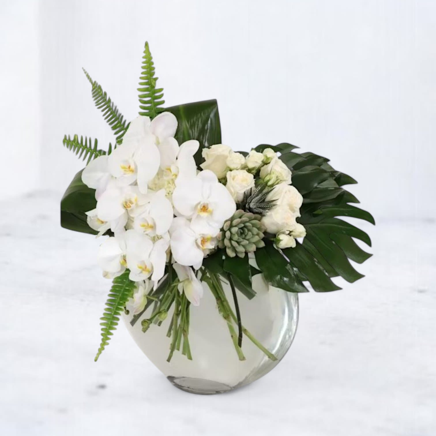 Ethereal Oasis - Experience the serene beauty of Ethereal Oasis, a modern and sophisticated arrangement that exudes tranquility and grace. Featuring cascading white orchids, delicate white spray roses, hydrangea, and a touch of lush greenery, this design is both minimalist and luxurious. A unique addition of succulents enhances its contemporary appeal, while the sleek glass vase completes the look with understated elegance. Perfect for special celebrations, condolences, or adding a touch of serenity to any space, this arrangement speaks the language of refined beauty and peace. Let Ethereal Oasis be your statement of timeless sophistication.