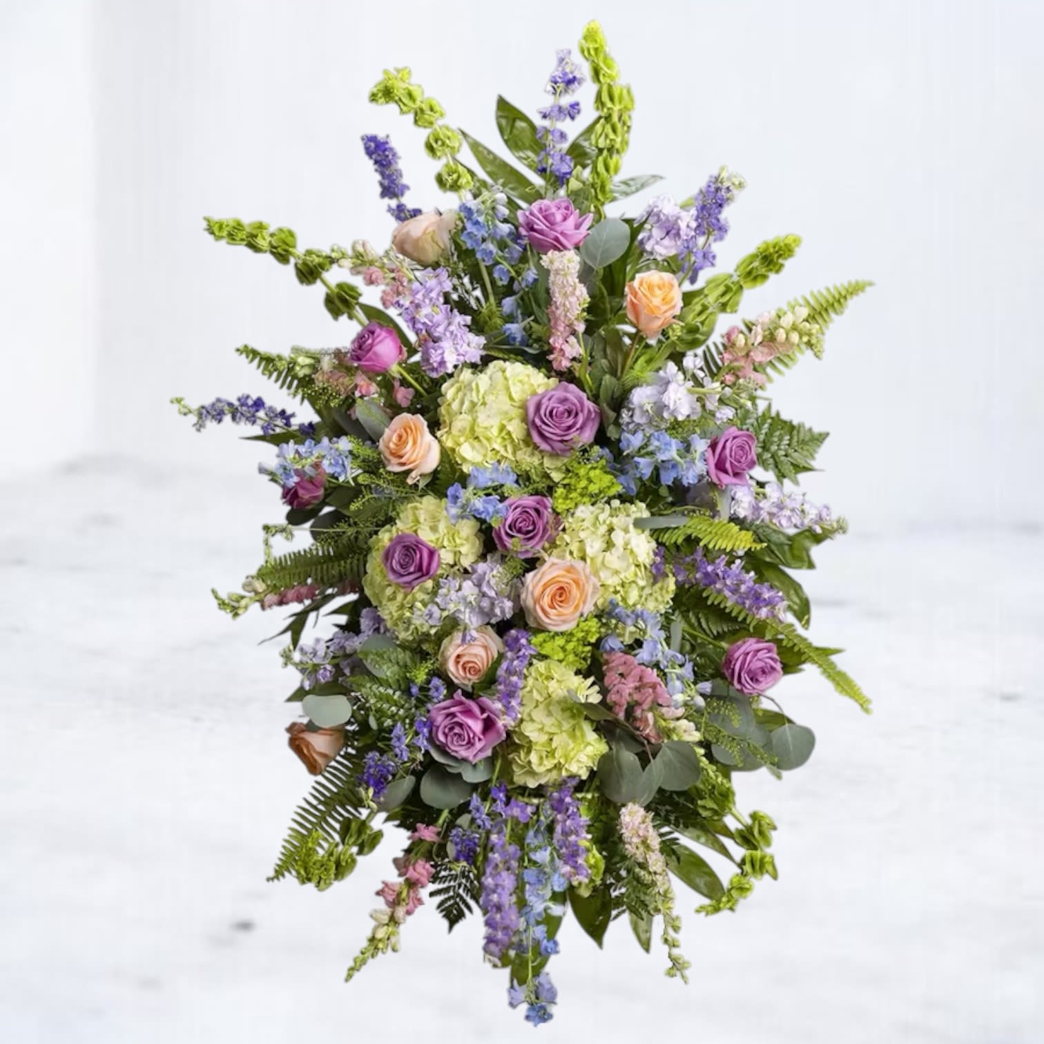 Standing Tribute - The Standing Tribute offers a soothing expression of love and remembrance. Featuring a harmonious blend of soft lavender roses, peach blooms, lush hydrangeas, and accents of delicate blue delphiniums, this arrangement embodies serenity and beauty. Cascading greenery adds a natural touch, while its gentle color palette provides a comforting tribute. Perfect for celebrating a life filled with grace and cherished memories, this standing spray brings an elegant and heartfelt presence to any memorial or service.  Easels are included with all standing sprays for effortless display at services. 
