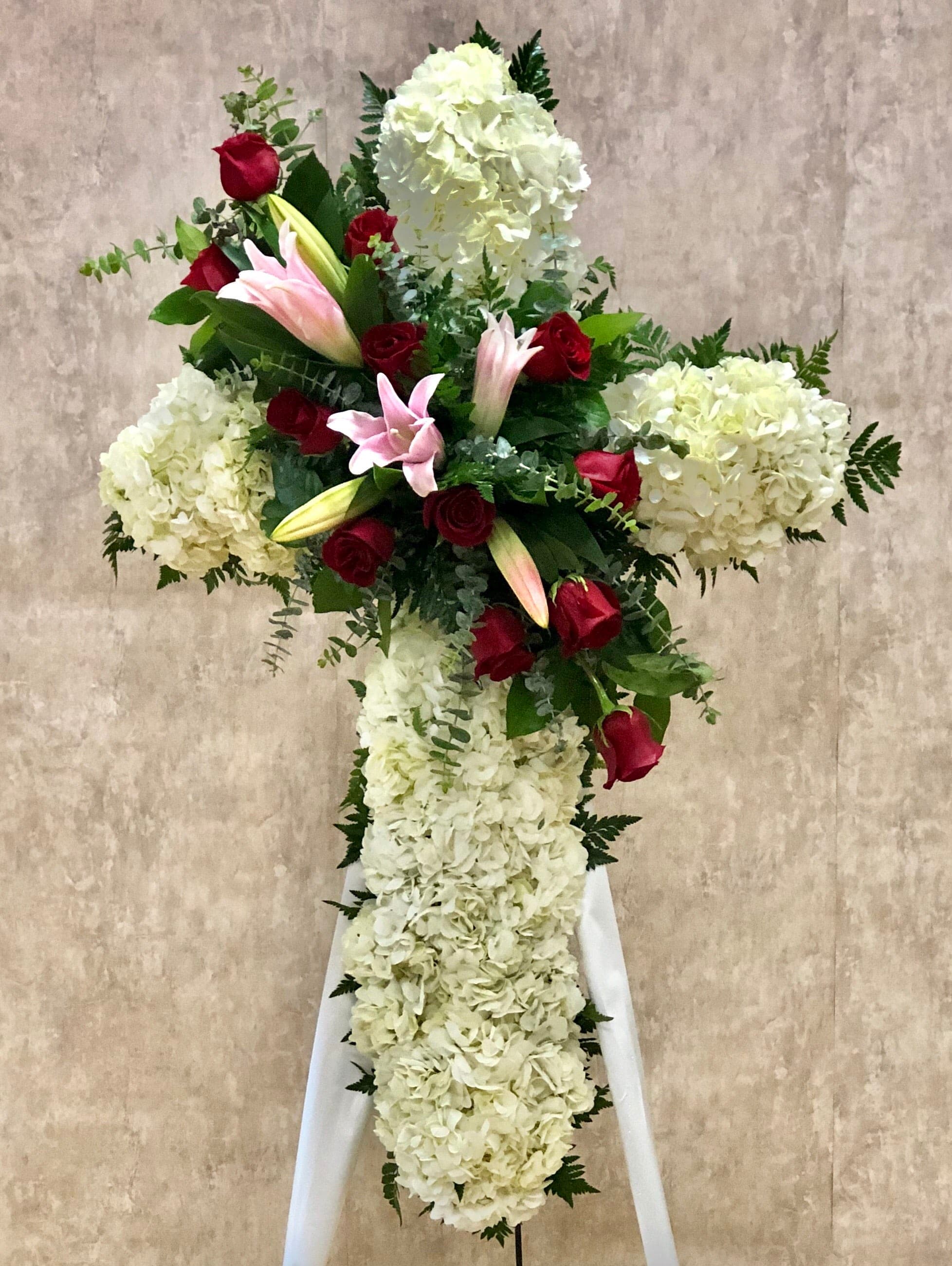 Eternal Peace Standing Spray (DB) - Honor a loved one with a meaningful tribute by choosing our standing cross spray. Meticulously crafted, this exceptional arrangement features white hydrangeas, pink lilies, and red roses, all arranged in the shape of a cross. It serves as a symbol of faith, love, and remembrance, holding a special significance in commemorating a life well-lived.