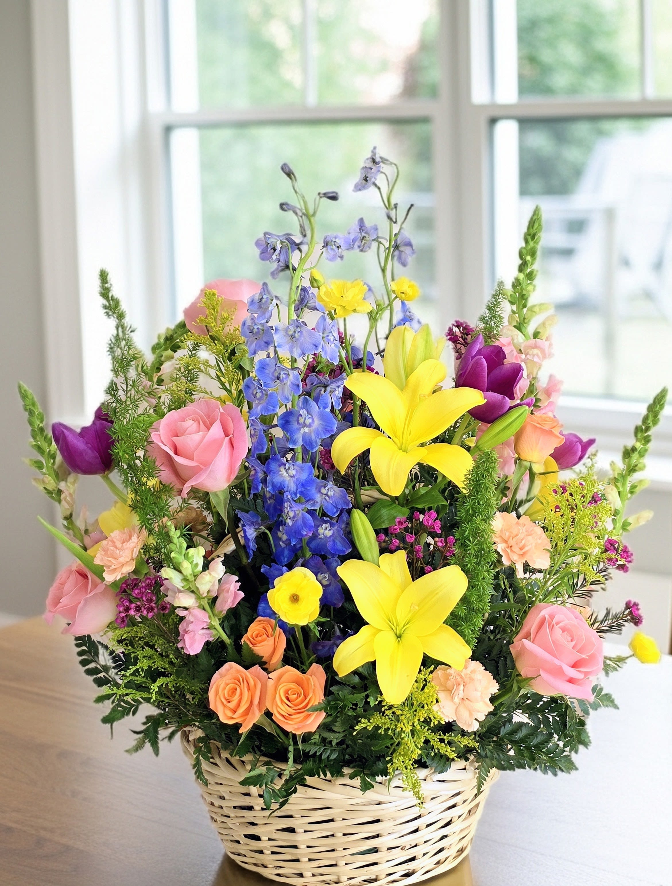Premium Spring Basket - Sunlit yellow lilies and sky-blue delphinium rise above a lush mix of pink and peach roses in this premium spring basket, creating a bright, garden-fresh centerpiece perfect for a birthday bouquet, spring celebration, or a congratulations arrangement. The upright, abundant silhouette and multicolor palette immediately read as cheerful and celebratory-the kind of gift that makes the person you're thinking of feel noticed and genuinely celebrated. Ideal for moms, friends, or colleagues marking spring milestones, new jobs, or special birthdays, this arrangement arrives ready to display in its wicker basket-no vase needed. For best enjoyment, keep blooms in a cool spot away from direct heat and refresh water every 3 days. Seasonal availability may vary, so order early to secure spring color.