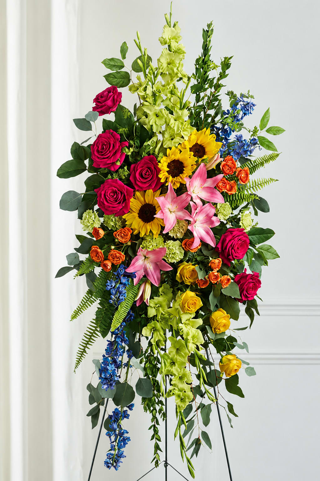 Celebration of Life Standing Spray - A vibrant and uplifting standing spray designed with a beautiful garden-inspired mix of bold and cheerful blooms. This elegant tribute features rich hot pink roses, radiant sunflowers, soft pink lilies, yellow spray roses, orange carnations, blue delphinium, and cascading green snapdragons accented with lush seasonal greenery and flowing foliage. Arranged on a standing easel with a natural cascading shape, the design symbolizes love, warmth, joy, remembrance, and a life beautifully celebrated. Its colorful palette offers a heartfelt and comforting expression of sympathy while honoring a vibrant spirit and cherished memories.