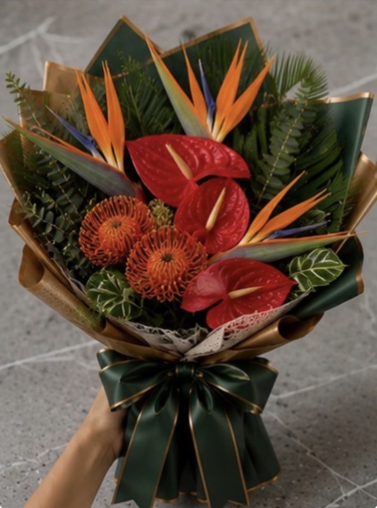 Tropical Luxe Wrapped Bouquet - Glossy red anthuriums and bright orange bird of paradise take center stage in this Tropical Luxe wrapped bouquet, an exotic flower arrangement perfect for a birthday celebration, anniversary, or any festive gifting moment. The upright cluster of dramatic tropical blooms reads as bold and celebratory - the kind of bouquet that makes the person you're thinking of feel seen and delighted the moment it arrives.  Dark green foliage and broad leaves form a lush backdrop that intensifies the saturated reds and oranges while hints of purple add depth to the palette. Wrapped in deep green and metallic gold decorative paper and finished with a large satin ribbon, the presentation is polished and formal-ideal for milestone events or as a standout statement arrangement. The compact, upright build makes it easy to display in a vase or as a striking wrapped gift; treat the stems with fresh water on arrival and keep them in a cool spot away from direct sun to extend their tropical beauty.