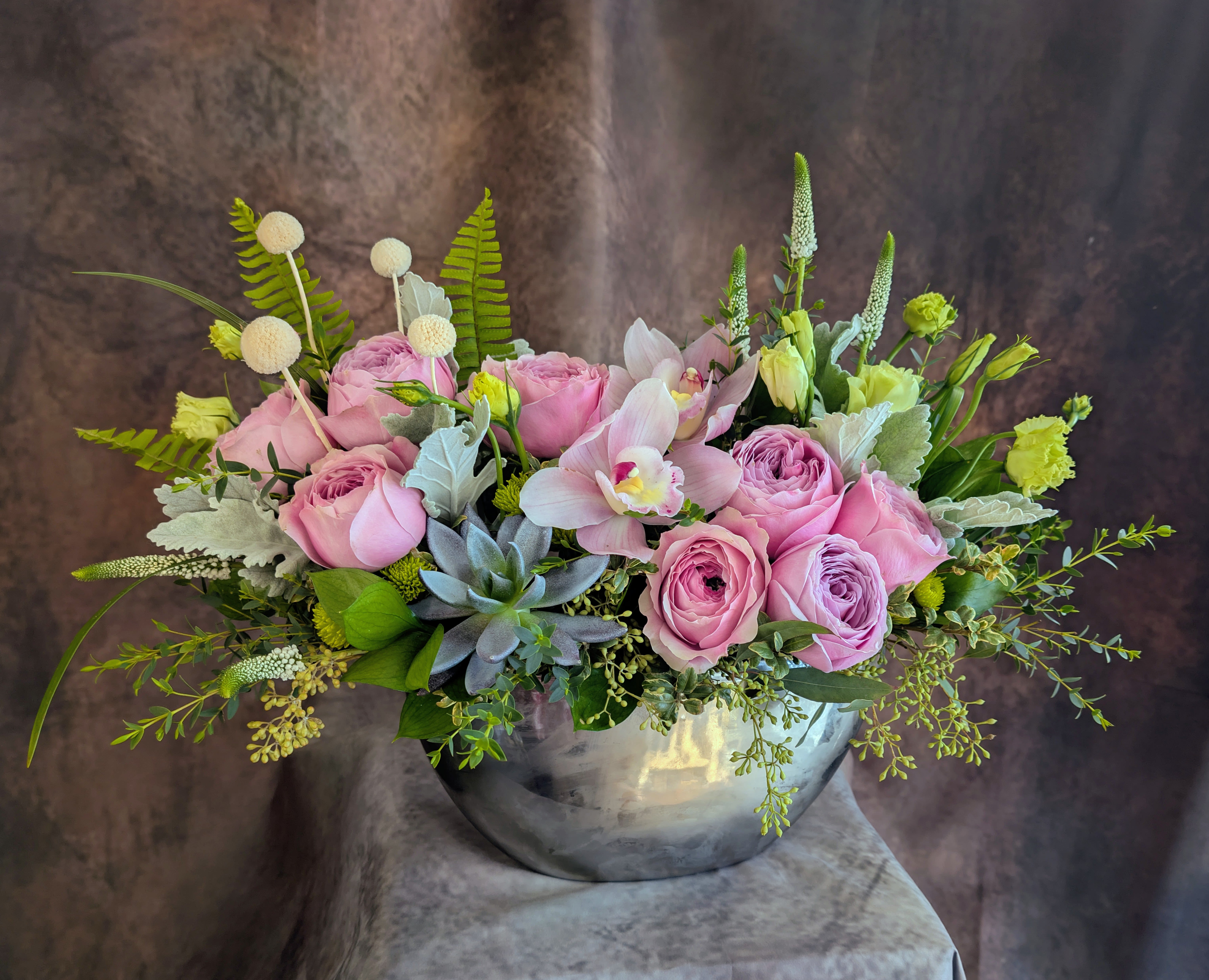 Paloma - Soft, luminous, and effortlessly refined, Paloma is a garden-style floral composition designed with a sense of lightness and natural movement. This piece brings together blush garden roses, delicate ranunculus, and elegant cymbidium orchids in a layered, textural arrangement that feels both modern and organic.  Accents of lisianthus, veronica, and seasonal greenery create depth and flow, while a sculptural succulent adds a subtle botanical contrast. Designed in a sleek silver vessel, Paloma balances softness with structure—offering a clean, elevated look with a relaxed, airy feel.  Thoughtfully composed with a focus on texture and form, this arrangement is ideal for Mother’s Day, birthdays, or meaningful everyday gifting. Its refined palette and natural movement make it a striking centerpiece without feeling overly formal.  Available for flower delivery in Las Vegas, Henderson, Anthem, MacDonald Ranch, and Inspirada, exclusively from Windmill Floral Expressions, a boutique florist known for artful, garden-inspired design.