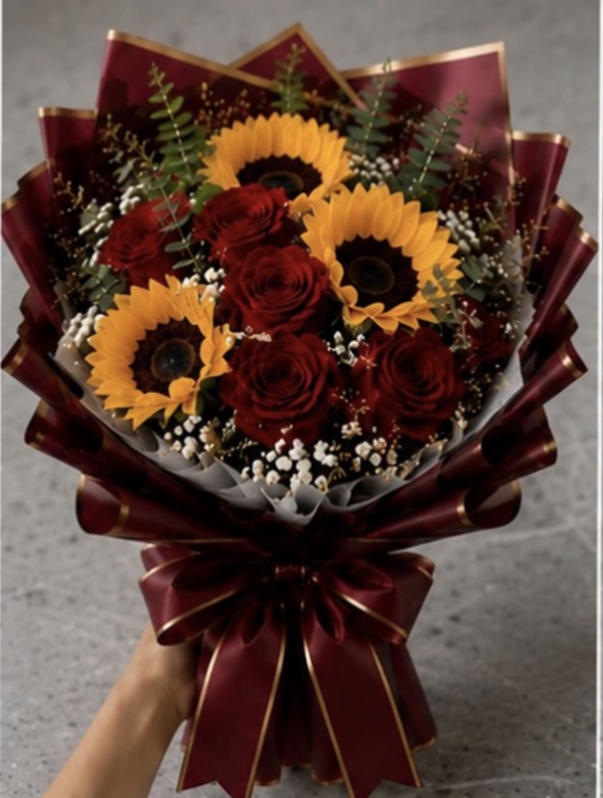 Sunflower and Rose Wrapped Bouquet  - Bold red roses paired with sunny yellow sunflowers create a striking birthday bouquet or anniversary gift that reads both romantic and celebratory. The contrast of deep crimson and warm gold makes a confident visual statement-perfect for someone you want to surprise and delight on a special day or to mark a meaningful milestone like an anniversary or congratulations moment.  Bright sunflowers and long-stemmed red roses are accented with small white filler blooms and airy greenery for texture and softness around the focal flowers. Wrapped in burgundy decorative paper with gold edging and finished with a large matching ribbon, this hand-tied bouquet arrives ready to present and makes an instant impression. Choose this wrapped bouquet when you want her to feel seen and celebrated; stems are arranged for easy vase placement and simple care-trim the stems, change the water every couple of days, and keep away from direct heat to prolong blooms.