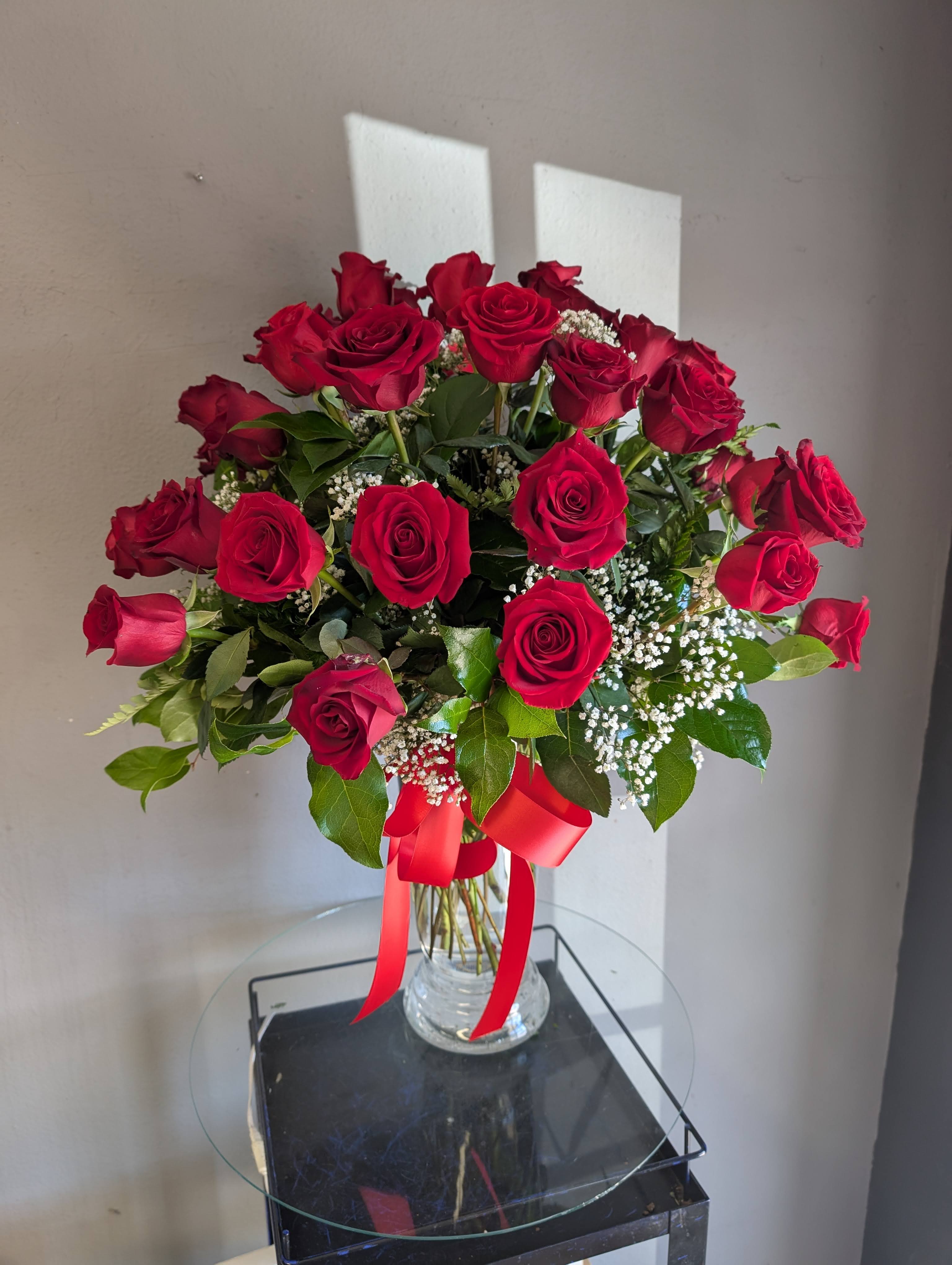 Tall arrangement of red roses with white filler in a clear glass vase tied with a red ribbon.