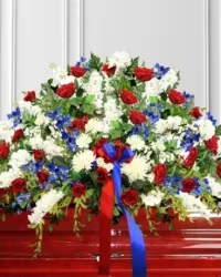 Patriotic Remembrance Casket Spray - A dignified and elegant casket spray designed in a classic red, white, and blue color palette to honor a cherished life. This beautiful tribute features red roses, white cushion mums, blue delphinium, white stock, and white alstroemeria, thoughtfully arranged with fresh greenery to create a full and graceful display.  Finished with a red and blue-ribbon bow, this arrangement symbolizes love, honor, and remembrance, offering a heartfelt and respectful tribute for a funeral or memorial service.