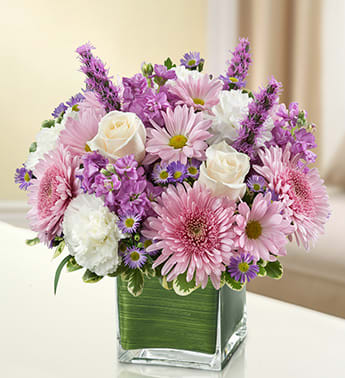 Pastel pink and lavender mixed flower arrangement in a square glass vase lined with green leaves