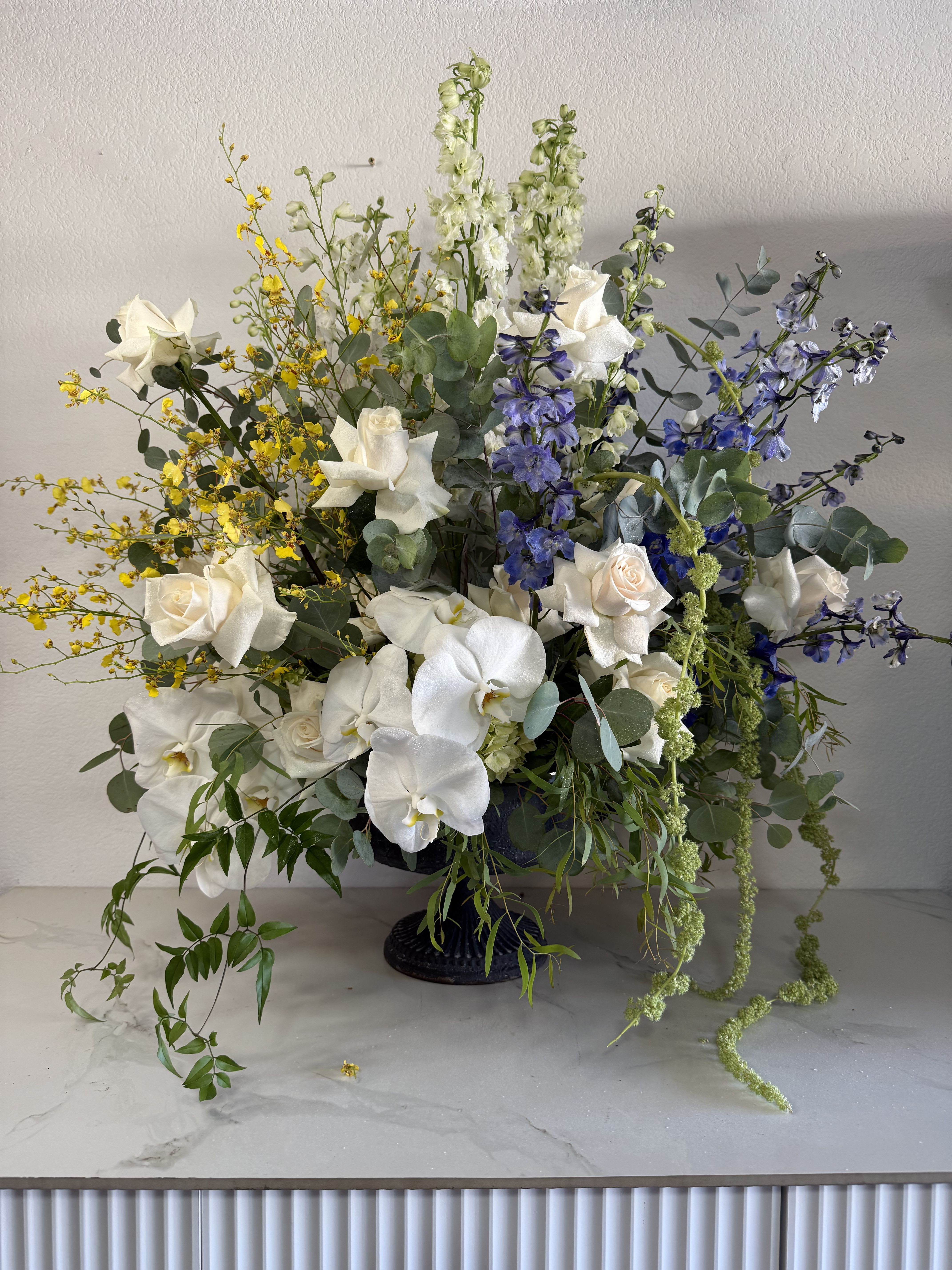 Ivory &amp; Indigo - White phalaenopsis orchids and white roses take center stage in this large, airy flower arrangement-an elegant flower arrangement perfect for a wedding, sympathy tribute, or any moment that calls for refined presence. Tall indigo-blue delphinium spikes lift the design, while fine yellow oncidium blooms add sunny contrast so the recipient feels comforted and celebrated at the same time.  Set in a dark pedestal vase, the composition spreads widely with cascading eucalyptus-like foliage and trailing greenery that soften the structure into a loose, garden-style silhouette. The white-and-blue base reads cool and sophisticated, the yellow accents bring warmth, and the mix of orchids, roses, delphinium, and complementary stems creates texture and graceful movement across a generous scale.  Ideal as a formal decorative centerpiece or a meaningful gift to mark a milestone, this arrangement suits upscale events as readily as bedside sympathy displays. Measures and vase details are reflected by the large pedestal presentation-please contact us for delivery timing or specific size requests to ensure it arrives fresh and properly styled.
