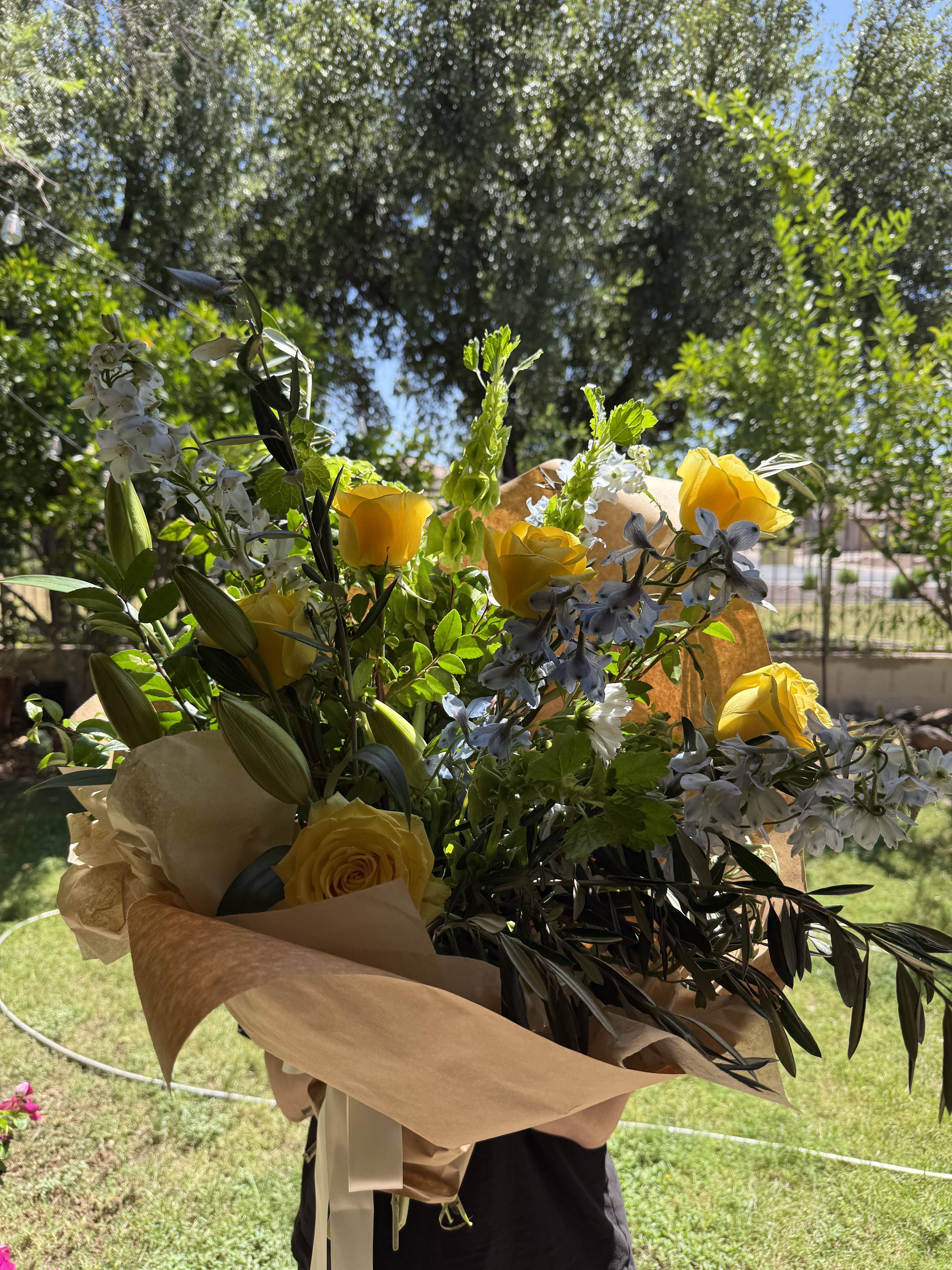 Yellow rose wrapped bouquet - Bright yellow roses take center stage in this hand-tied flower arrangement, sending an immediate note of warmth and celebration-perfect as a birthday bouquet, a thank-you gift, or a springtime surprise for someone you want to brighten. The combination of yellow roses with white lily blooms and pale blue delphinium-like spikes creates a joyful, fresh contrast that reads both cheerful and thoughtfully composed for everyday celebrations or congratulations.  The bouquet is arranged loose and airy with long stems for a garden-picked look, wrapped in layered tan and peach-toned brown paper and finished with a pale ribbon for a natural, rustic presentation. You'll find sunny yellow roses, crisp white lilies, cool blue delphinium-like accents, and touches of green foliage that add texture and movement throughout the arrangement.  This is the kind of bouquet that makes the recipient feel genuinely celebrated-bright, uplifting, and slightly informal so it works for friends, family, or coworkers. Stems are left long for a generous feel; if you prefer a shorter vase-ready style, you can trim before arranging. Order for same-week delivery to capture its fresh, seasonal charm.