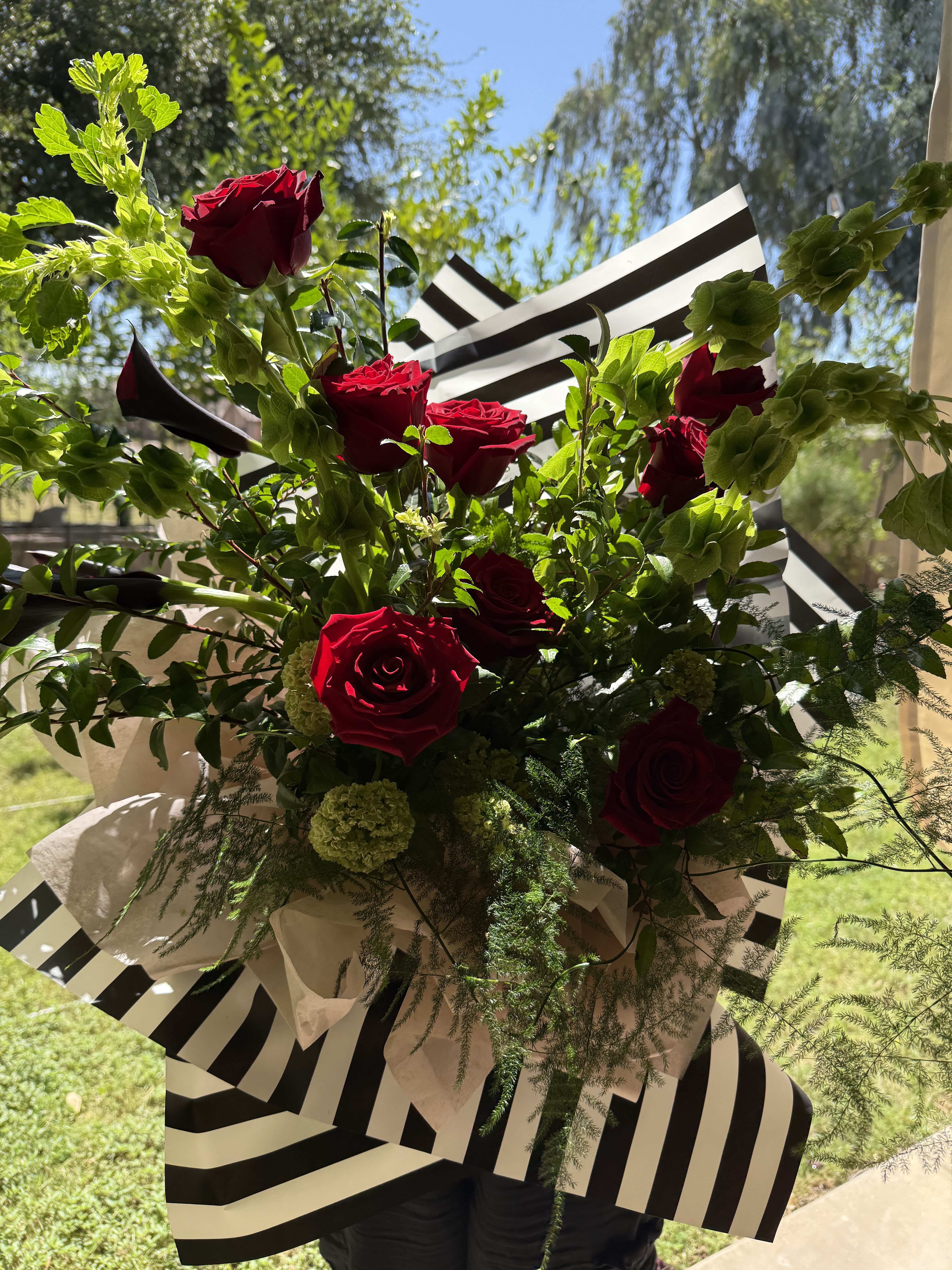 Red rose bouqet - Deep, velvety red roses arranged with airy greenery and mini calla lilies create a bold, modern romantic statement - perfect as a birthday, anniversary, or "thinking of you" bouquet. The hand-held arrangement reads celebratory and intimate at once, making the recipient feel truly seen whether you're marking a summer milestone, a spring celebration, or simply sending love and romance just because.  Lush Bells of Ireland and soft white accents lift the composition, adding texture and height against the saturated red blooms and vivid green foliage. The bouquet is wrapped in striking black-and-white striped and decorative paper for a contemporary contrast that makes the color pop and gives a polished presentation suitable for Mother's Day, Women's Day, Valentine's Day, or congrats moments.  Ideal for someone who appreciates dramatic, elegant florals, this arrangement arrives ready to display or carry to a celebration. The layered, full shape holds up beautifully in a vase; trim stems and refresh water daily to extend vase life and enjoy the scent and color for as long as possible.