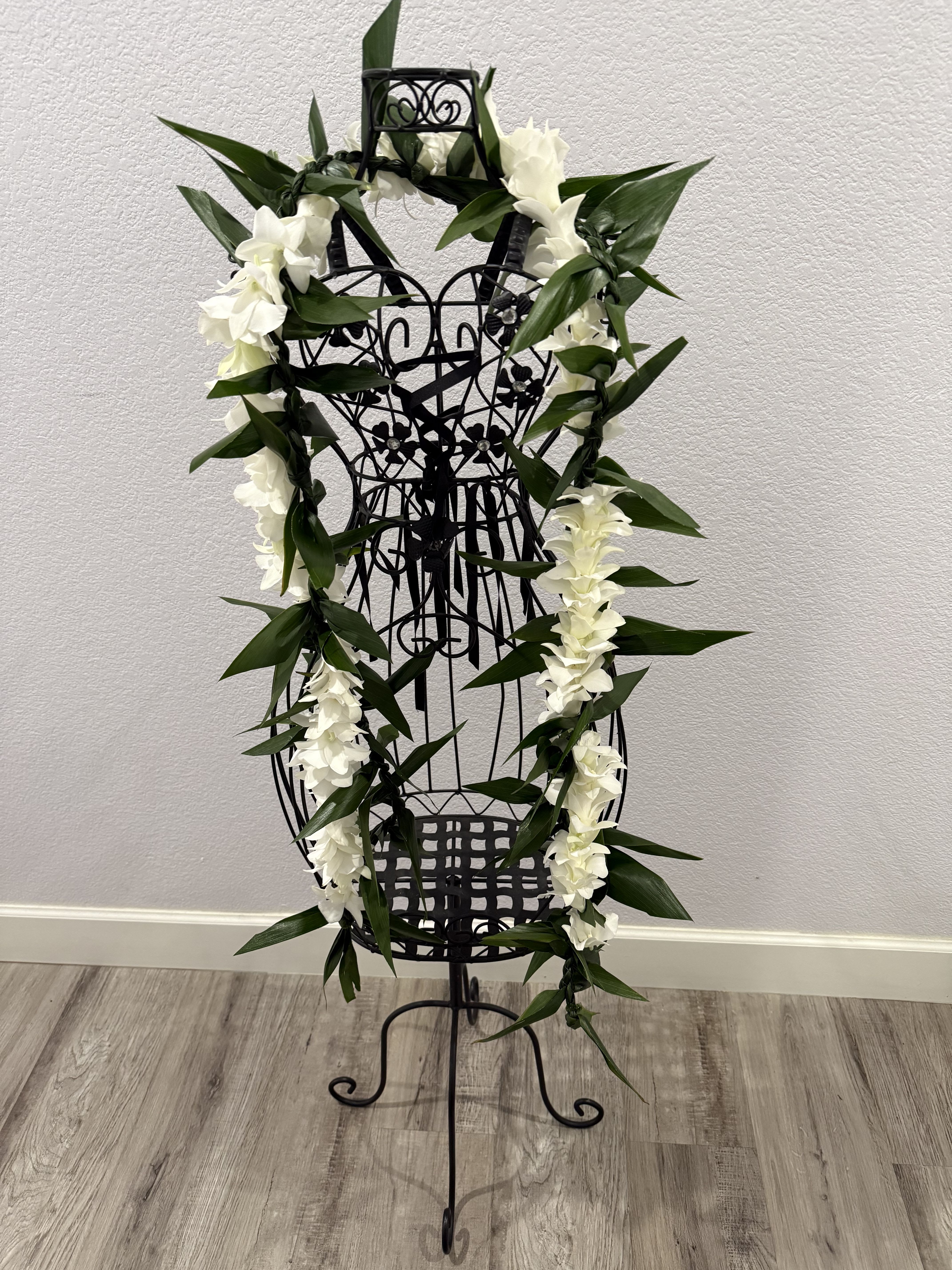Orchid and ti leaf lei - A long white orchid and ti leaf lei draped in a vertical display makes a quiet powerful floral statement, perfect as a ceremonial flowers. The white blooms read pure and serene against broad dark green leaves suits for weddings or graduation  The design pairs a long, garland-style lei with deep green ti leaves to create strong contrast and a graceful vertical line.  Choose this arrangement when you want the person you're honoring to feel respected and seen-whether marking a life remembered or lending formality to a wedding ceremony. This piece is hand made in house, call us with instructions