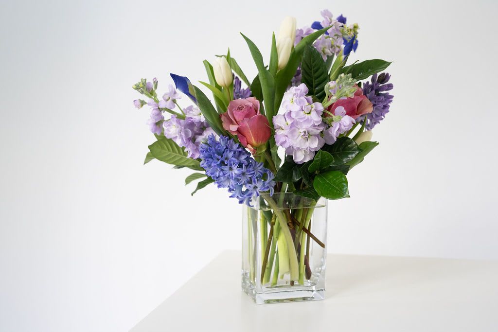 Mixed bouquet of pink roses, purple stock, blue blooms, and white tulips in a clear glass vase