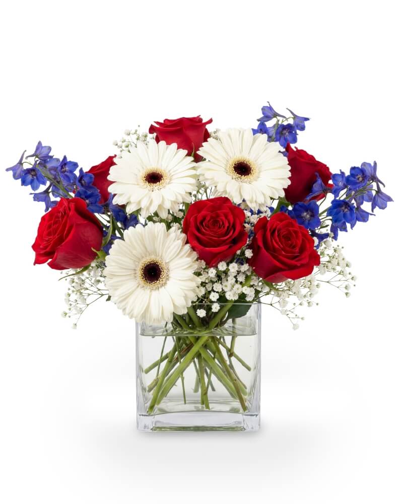 Liberty Glow - Bright as a summer sunrise and bold as the American spirit, Liberty Glow captures the heart of July 4th in a modern, joyful display. Vivid red roses rise proudly among crisp white daisies and airy sprigs of baby’s breath, while bursts of blue delphinium give a fireworks-like flare. Designed in a sleek glass cube vase with visible stems and water, this arrangement beams with patriotic charm and a touch of elegance—perfect for celebrating freedom, family, and the beauty of summer.