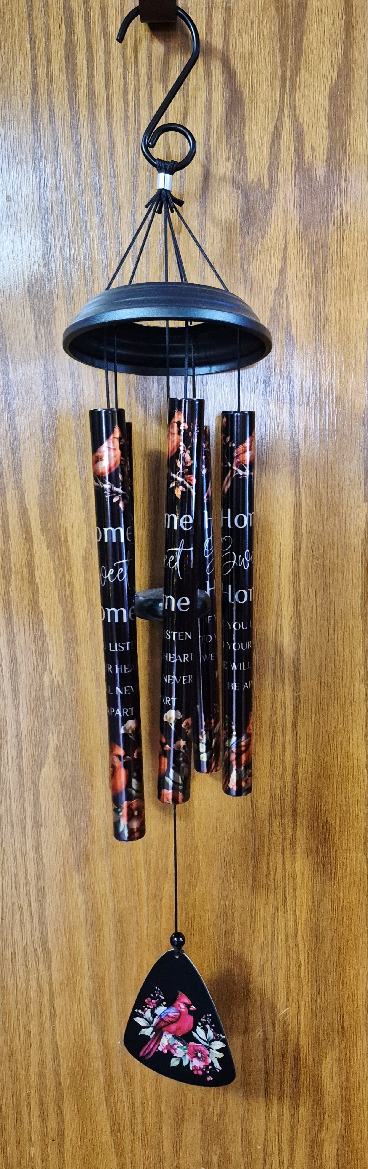 Home Sweet Home Wind Chime - A memorial wind chime crafted to offer comfort and a gentle, lasting tribute-perfect for sympathy and funeral gestures. The Home Sweet Home wind chime pairs somber black metal tubes printed with floral graphics and remembrance text with a soft pink bird pendant, creating a visual and sonic keepsake that helps the person you're thinking of feel remembered and soothed.  Hand-tuned for clear, resonant tones, the chime measures 30" from the top gathering ring to the bottom of the wind catcher and includes a 2" hook for easy hanging. The pendant's pink blossom and bird detail adds a tender contrast to the dark finish, while the printed message-"Home Sweet Home / If You Listen To Your Heart We Will Never Be Apart"-offers a heartfelt sentiment without words spoken aloud.  For a formal presentation at a service or graveside, pair the chime with the optional wire easel stand (Deluxe option includes the stand and its $15 charge) to display the piece upright. This keepsake is suitable year-round as a thoughtful remembrance gift and requires only an outdoor or sheltered spot where breezes can activate its calming melody.