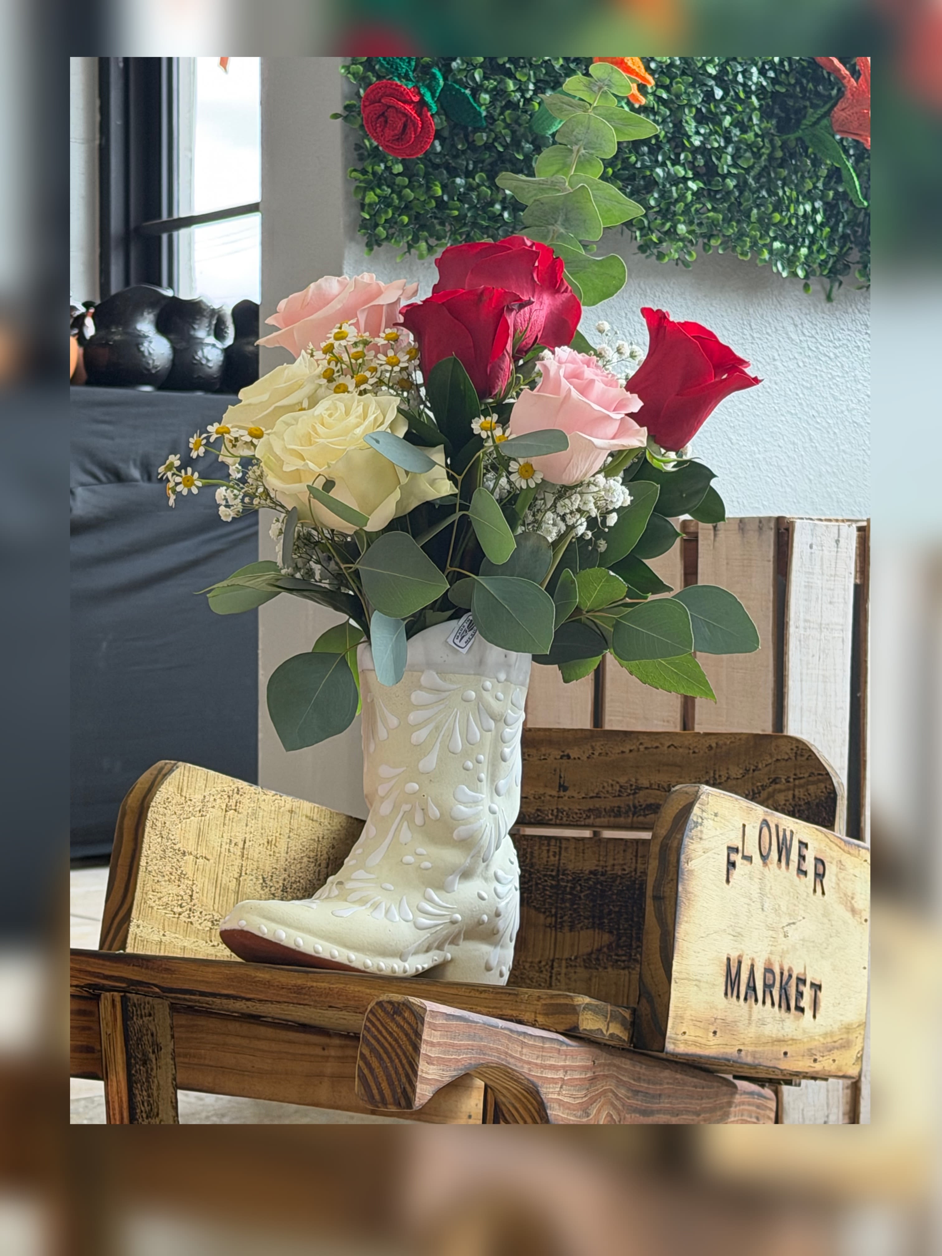 Country Delight - Red and rosy charm makes this Mother's Day bouquet feel like a warm country hug - a flower arrangement built around clustered red, blush-pink, and creamy ivory roses that say "you're loved" for Mother's Day, birthdays, anniversaries, or a thoughtful just-because. Small white daisy-like filler flowers and airy baby's breath create a soft, romantic halo around the roses so the person you're thinking of will feel genuinely seen and celebrated.  The flowers are gathered in a whimsical white ceramic boot vase, nestled with rounded eucalyptus stems that add cool gray-green contrast and gentle height. The compact, rounded silhouette and textured mix of roses, delicate daisies, and airy greenery give a cheerful, handcrafted look; shown tucked into a rustic wooden crate or sitting on a wooden bench, it reads as a charming keepsake piece for a kitchen table or bedside.    Perfect for anyone who loves country-inspired style, this arrangement arrives ready to display. Keep water fresh and trim stems every few days to extend vase life.