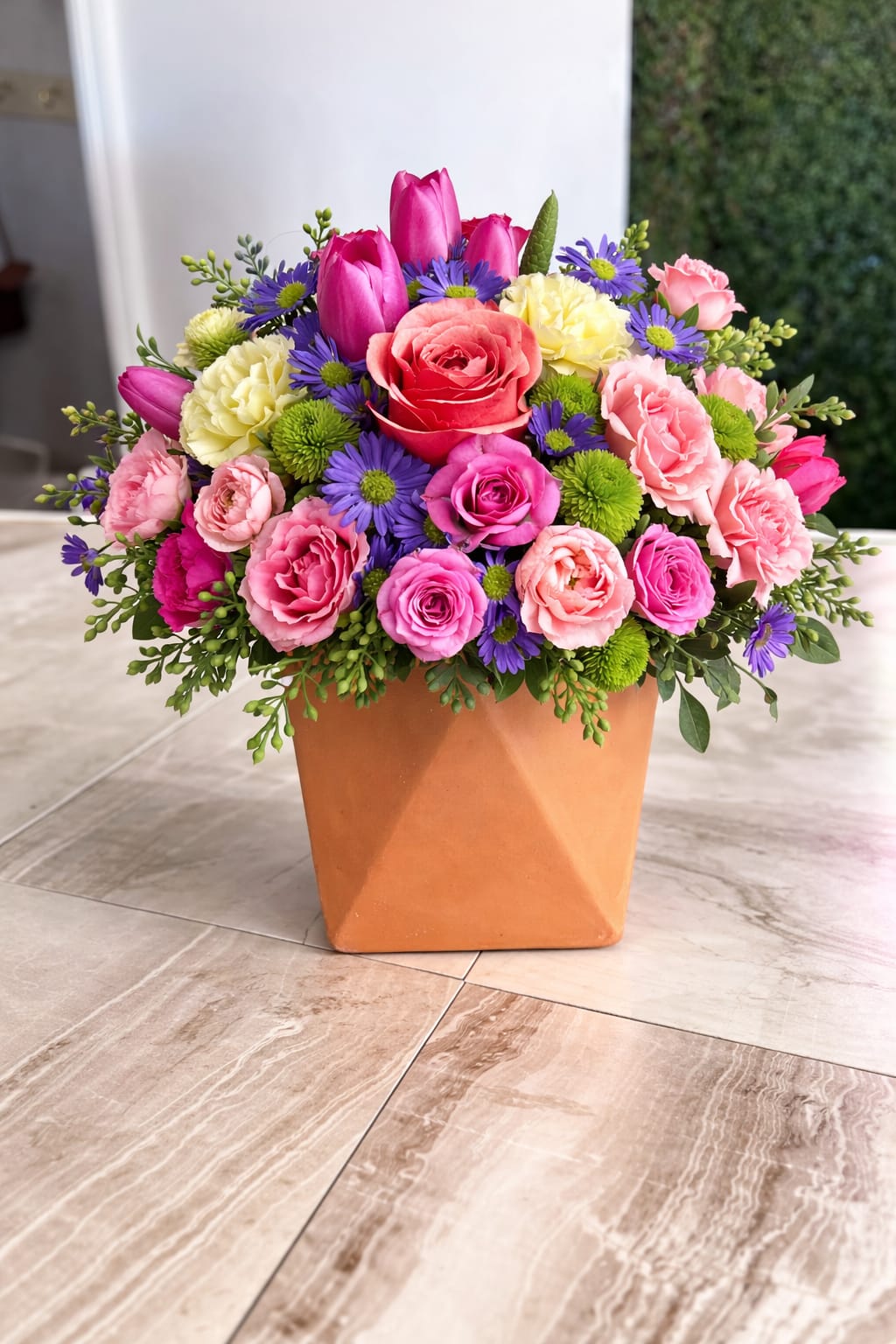 Bright mixed arrangement of pink roses, tulips and purple daisies in a terracotta cube vase