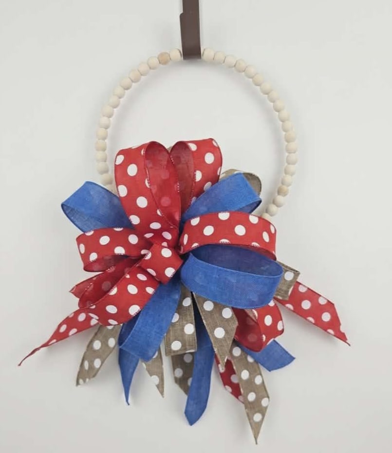 Red, White and Blue Beaded Wreath  - A bold ribbon-adorned hanging ring that reads as festive home decor-perfect for patriotic displays or a Memorial Day, Fourth of July, or summer front-door accent. This flower arrangement alternative makes the person you're celebrating feel noticed with its layered red, white and blue theme and eye-catching bow instead of traditional blooms.  The circular beaded frame is finished with a large, layered bow made from red polka-dot ribbon, solid blue ribbon, and tan polka-dot ribbon so the focus stays on texture and color contrast. The neutral background of the wreath keeps attention on the playful mix of patterns and the sturdy beaded hanging ring and ribbon bow mean it's ready to display right out of the box.  Ideal for gifting to someone who loves handcrafted, Americana-style decor or for marking seasonal occasions on a porch, entryway, or event space.  Wreath is 10" around not measuring bow that is included.