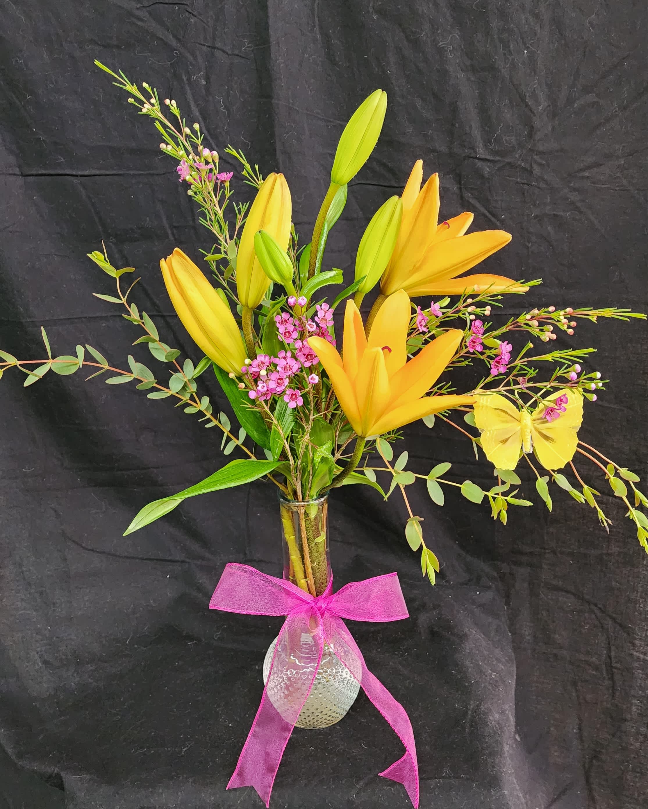 Yellow lilies in a glass vase with pink flowers and a pink ribbon