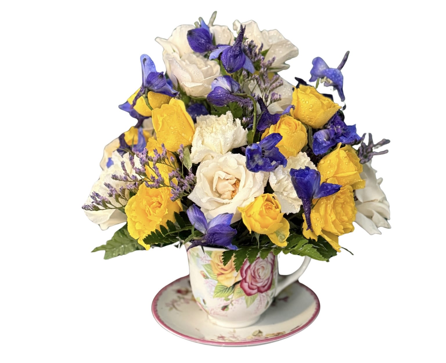 Flowers on tea cup - A sunny cluster of yellow and white roses nests in a decorative ceramic teacup on a matching saucer, creating a whimsical Mother's Day or birthday flower arrangement that feels like a hug in porcelain. This flower arrangement for Mom pairs the soft romance of pink accents with purple lisianthus-like blooms and tiny lavender fillers, so the person you're celebrating will feel brightened and truly seen.  Compact and rounded for easy placement on a bedside table or kitchen windowsill, the piece blends vintage-inspired charm with everyday freshness. Roses and lisianthus provide layered texture and scent, while the included ceramic teacup and saucer make the gift ready to display-perfect as a thank-you, spring celebration, or just-because surprise. Ordering is simple and the size is ideal for close-up enjoyment where she can admire the colors up close.