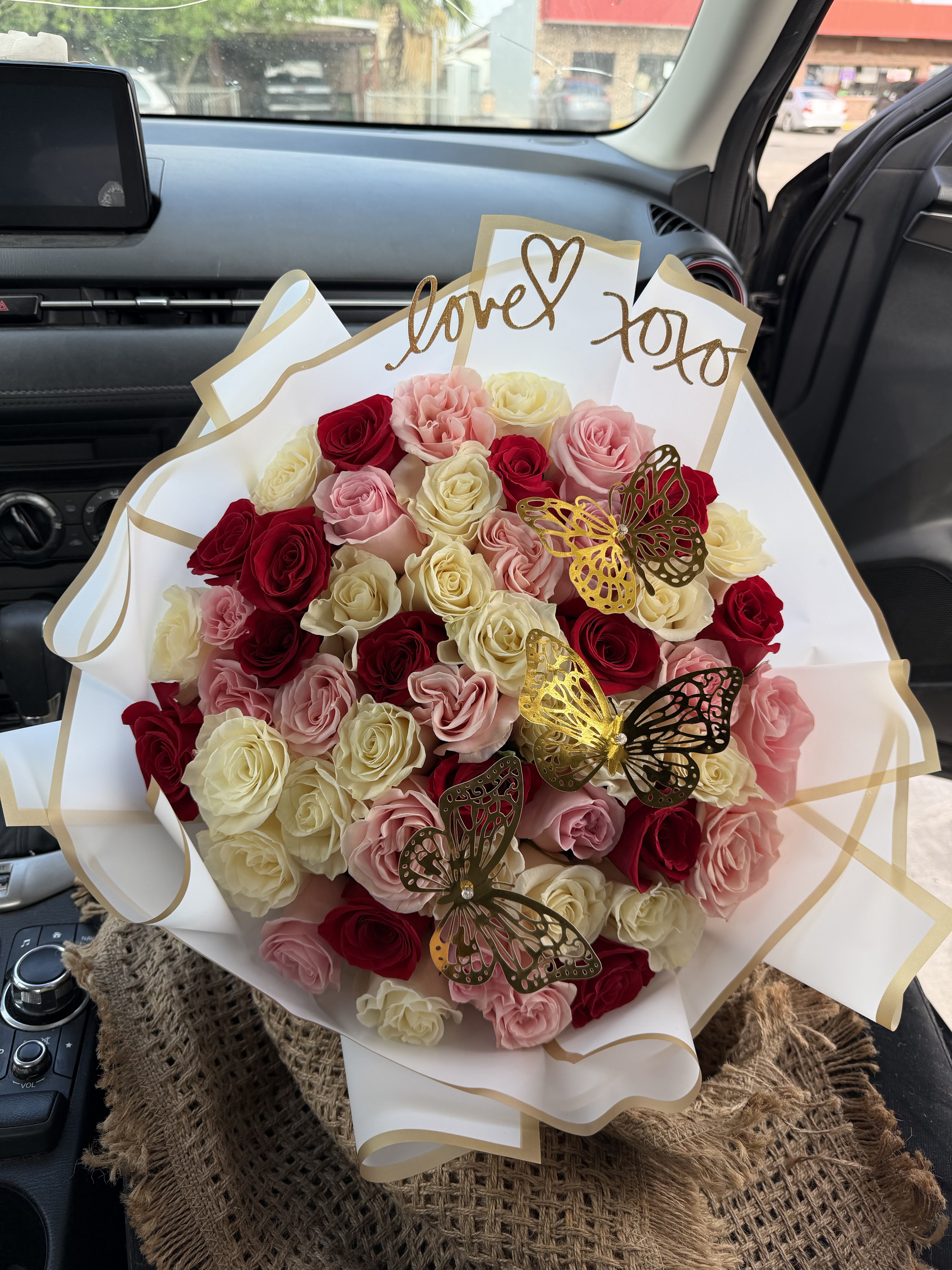 quadruple tri colored buchona para mama - A lush, rounded spray of roses in blush pink, deep red, creamy ivory and a touch of sunny yellow creates an immediate feeling of warmth-perfect as a birthday bouquet, anniversary roses, or a romantic Valentine's Day surprise for mamá. The tri-colored buchona para mama reads like a heartfelt celebration: the mix of soft pastels and saturated reds makes her feel both cherished and joyfully celebrated.  Tightly packed roses form a dense, hand-tied shape wrapped in crisp white paper with gold edging, trimmed with a gold ribbon for a gift-ready finish. Decorative metallic butterfly picks and a gold glitter script topper add playful, shimmering accents that catch the light and lift the arrangement's texture. The compact size and full presentation make it easy to place on a table or bedside without losing impact.  This arrangement is an ideal way to tell the person you're thinking of that she's loved-whether for Mother's Day, an anniversary, or just because. Care is simple: keep roses in fresh water, trim stems occasionally, and display away from direct heat to extend their life.