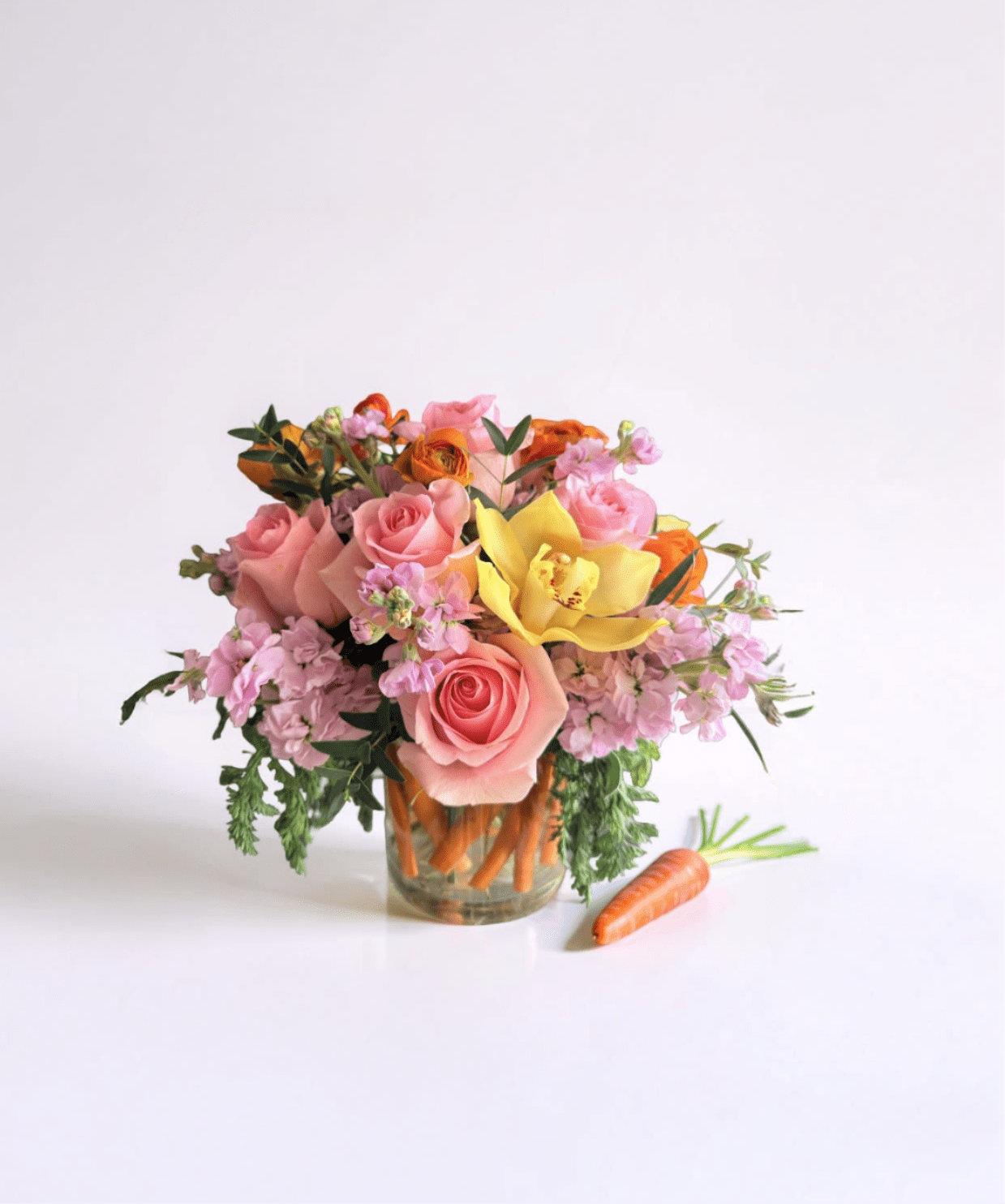 Short arrangement of pink roses, yellow orchid, and orange flowers in a carrot-filled glass vase with a carrot beside it