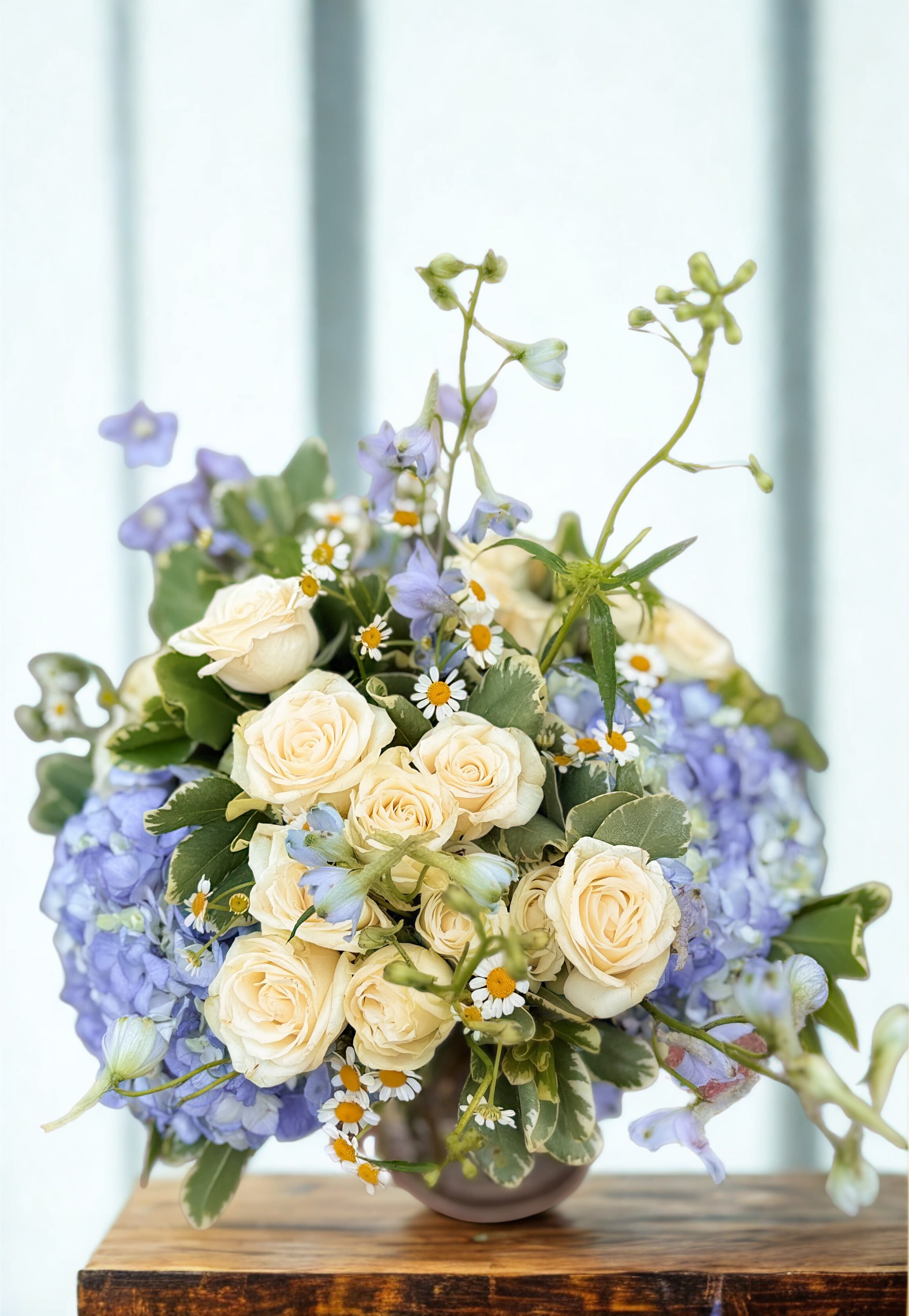 Compact arrangement of cream spray roses, lavender hydrangeas, and small daisies in a round vase.