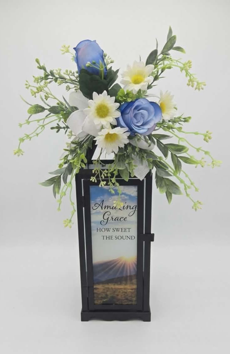 Amazing Grace Panoramic Lantern Lighted w/ Silk - Silk blue roses and white daisy-like blooms float above a black lantern in this sympathy flower arrangement, creating a calm, devotional display perfect for a memorial or remembrance setting. The piece reads as a heartfelt tribute-its printed "Amazing Grace" panel on the lantern front gives the arrangement a reverent focus that will comfort the person you're honoring.  Airy, asymmetrical stems and light green filler give the silk arrangement a softly lifted silhouette; textures of roses, daisies and assorted silk florals contrast gently with the lantern's clean metal and clear glass. The lantern is finished in powder-coated black and includes the printed memorial insert, with overall dimensions of approximately 4.75" wide x 15.5" high x 4.75" deep so it can stand on a table, shelf, or beside a graveside marker.  Designed to serve as a keepsake accent, this arrangement suits sympathy services, memorial corners, or ongoing remembrance at home-she'll feel that her loved one is being honored with quiet dignity. Because it's crafted with silk flowers and a durable metal lantern, it arrives ready to display and will maintain its respectful presence without special care.