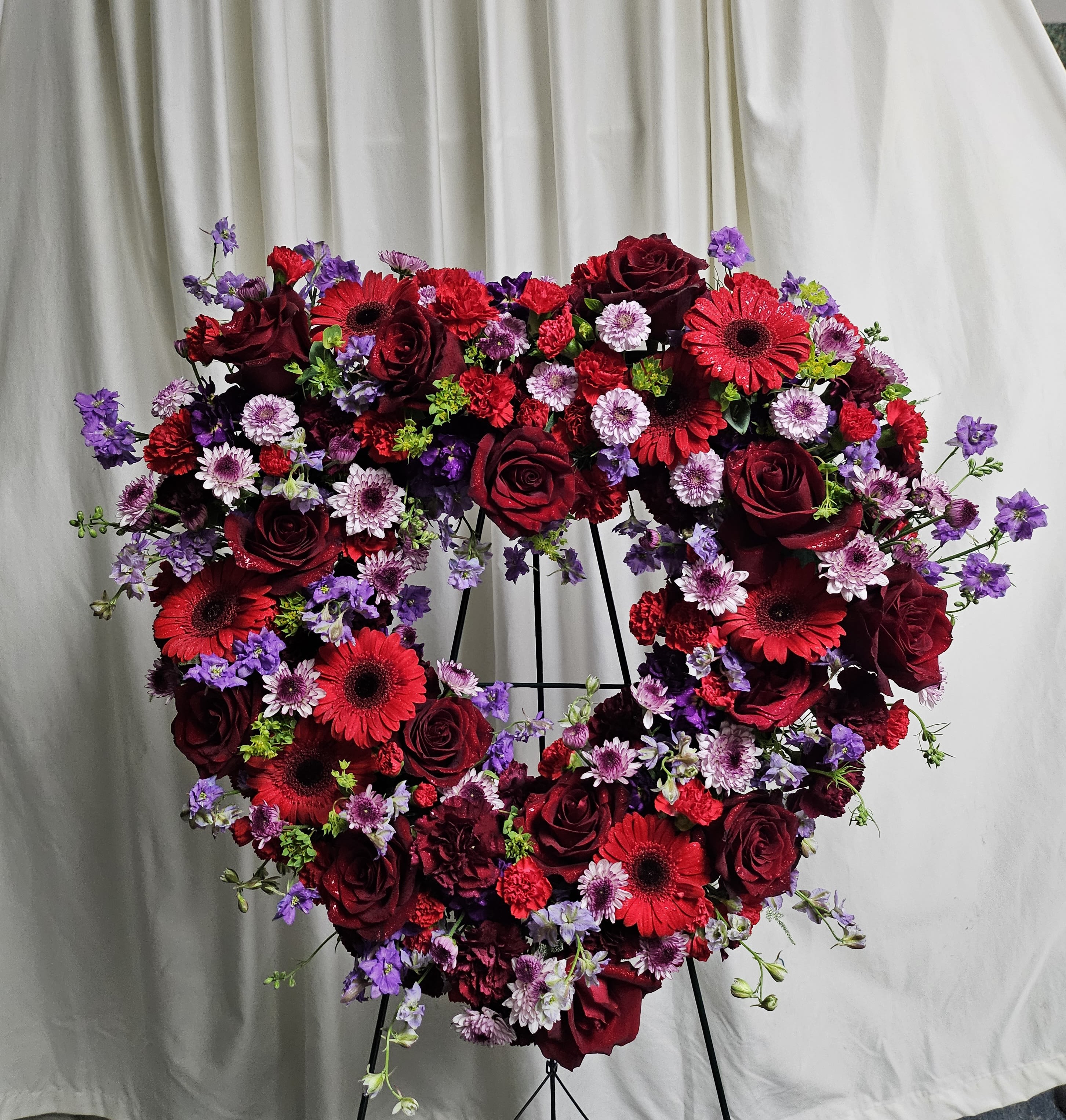 With Our Love - Full open heart with Roses, Gerbera daisies, mixed Carnations, light additions of button poms and larkspur. Colors may be substituted upon request and availability. 24" wide.
