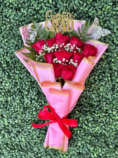 Pink Congratulations Flat Bouquet of Roses - A tight cluster of vivid red roses wrapped in soft pink paper with gold edging creates an instantly celebratory, romantic statement - perfect as a graduation bouquet or a birthday roses gift. The gold "Congrats Grad" and other celebratory toppers plus decorative butterfly picks make the arrangement read unmistakably milestone-ready, so the person you're celebrating will feel seen and special the moment they open it.  The bouquet pairs richly colored red roses with small white filler blooms and fern-like greenery for contrast and texture. Wrapped in pink paper, finished with a bright red ribbon and gold accents, it has a polished, gift-like presentation that reads both youthful and elegant. Sized as a hand-tied flat bouquet, it sits comfortably in the hand and is easy to carry to ceremonies or surprise visits.  Choose this arrangement for graduations, birthdays, anniversaries, or any moment you want to mark with warmth and flair - the toppers make the occasion clear while the classic roses convey affection. For best enjoyment, keep the stems in fresh water, trim them slightly before placing in a vase, and display out of direct sunlight to extend vase life.