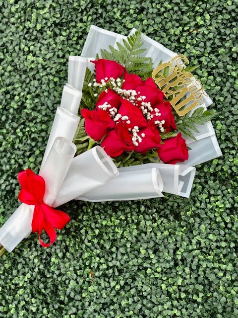 Congrats Grad White Flat Bouquet Ramo - Vivid red roses take center stage in this Congrats Grad bouquet, making a bold, celebratory statement perfect for graduation gifts or an anniversary-style congratulation. The tight cluster of crimson blooms against crisp white accents and fern-like greenery creates a striking red-and-white contrast that reads both romantic and triumphant - the kind of bouquet that makes the person you're honoring feel truly seen and celebrated.  Carefully wrapped in layered white paper and finished with a bright red ribbon, the arrangement arrives gift-ready with a polished, photographed-worthy look. Small white filler flowers weave through the roses for texture, while the translucent white wrap keeps the focus on the blooms. A gold "Congrats Grad" topper and decorative gold accent add a festive, personal touch that signals the occasion at a glance.  Send this to a recent graduate, a proud parent, or anyone reaching a milestone to convey warmth, admiration, and excitement. Size and care: wrapped bouquet-handle gently, keep stems in fresh water and trim an inch before placing in a vase to extend vase life.