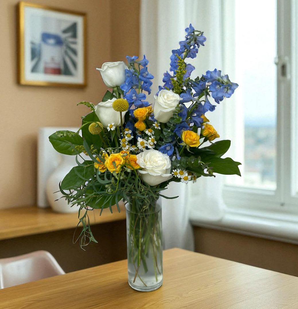 Tall arrangement of white roses, blue delphinium, and yellow blooms in a clear glass vase on a wooden table