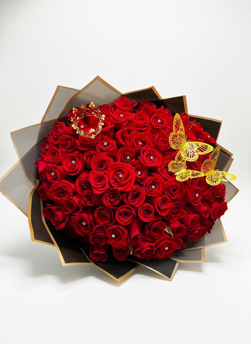 Ramo Buchon "Te Amo" - A dramatic, heart‑felt statement for anniversaries and romantic occasions: this tall floral arrangement brings deep red roses and a pink-and-white stargazer lily together to say "Te amo" with real presence. The composition reads romantic and celebratory-perfect for the person you love when you want them to feel cherished and seen.  The design layers a cluster of deep red roses above a showy stargazer lily, balanced by a pale hydrangea bloom, trailing burgundy amaranthus and glossy green foliage for texture and movement. A second presentation offers the roses tightly packed into a rounded dome, wrapped in black-and-gold decorative paper and finished with playful butterfly and jeweled picks for high-contrast drama. Both sit in a clear cylindrical glass vase and include decorative wrapping accents that elevate the presentation.  Ideal for anniversaries, Valentine's Day, or any moment when you want to make a bold romantic impression, this arrangement suits recipients who love classic roses with a modern, embellished twist. Vase-arranged for easy display; refresh water daily and recut stems every few days to keep blooms looking their best.