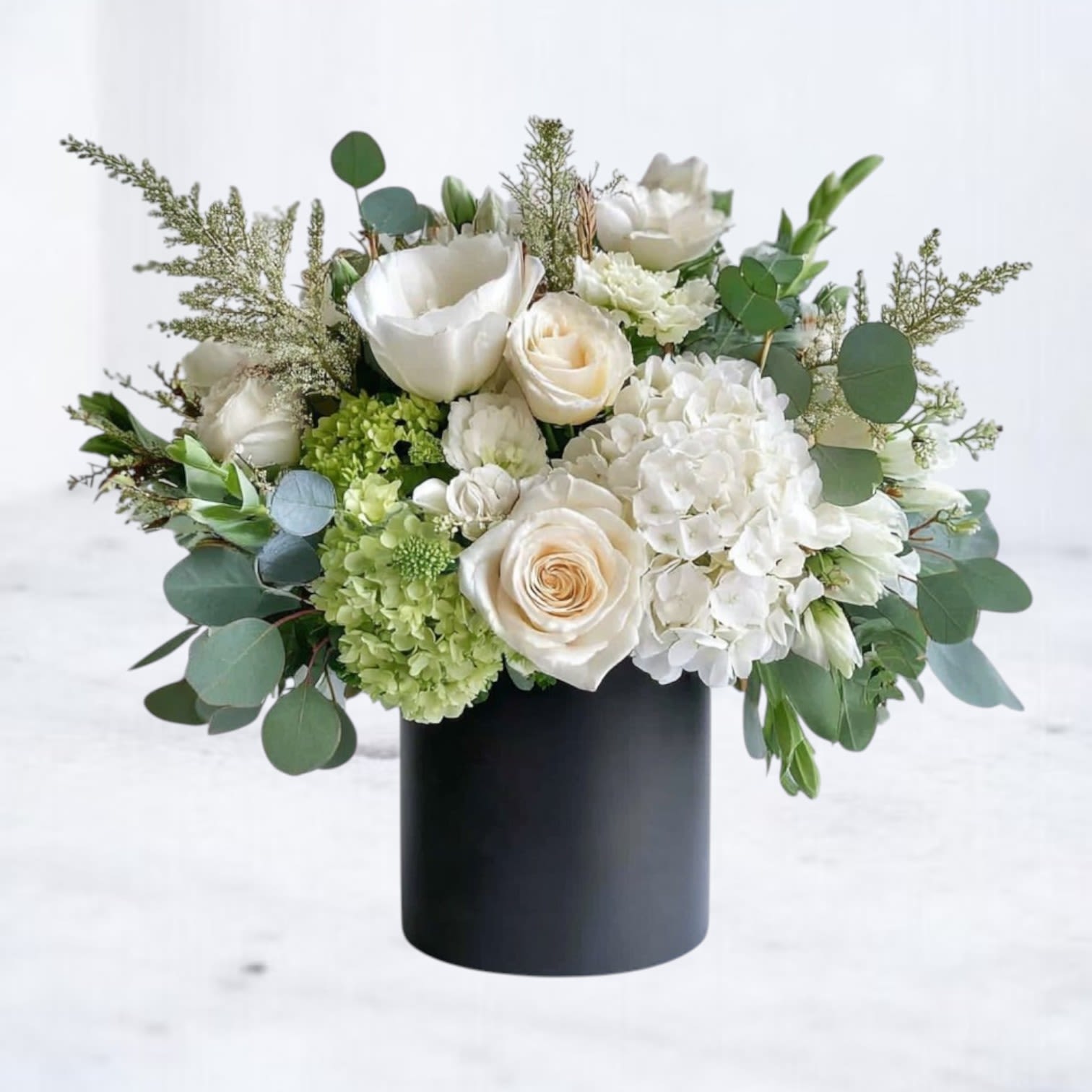 Pearl Essence - Pearl Essence is a sophisticated floral arrangement that radiates timeless elegance. Featuring lush ivory roses, delicate ranunculus, and soft lisianthus, this bouquet is accented with fresh greenery and delicate filler blooms. Presented in a sleek matte black cylinder vase, it offers a stunning contrast that enhances the natural beauty of the flowers. Perfect for sympathy gestures, or sophisticated home décor, Pearl Essence brings a serene and graceful ambiance to any setting.  Order this luxurious bouquet for delivery in Houston and surrounding areas to express your sentiments with classic beauty.