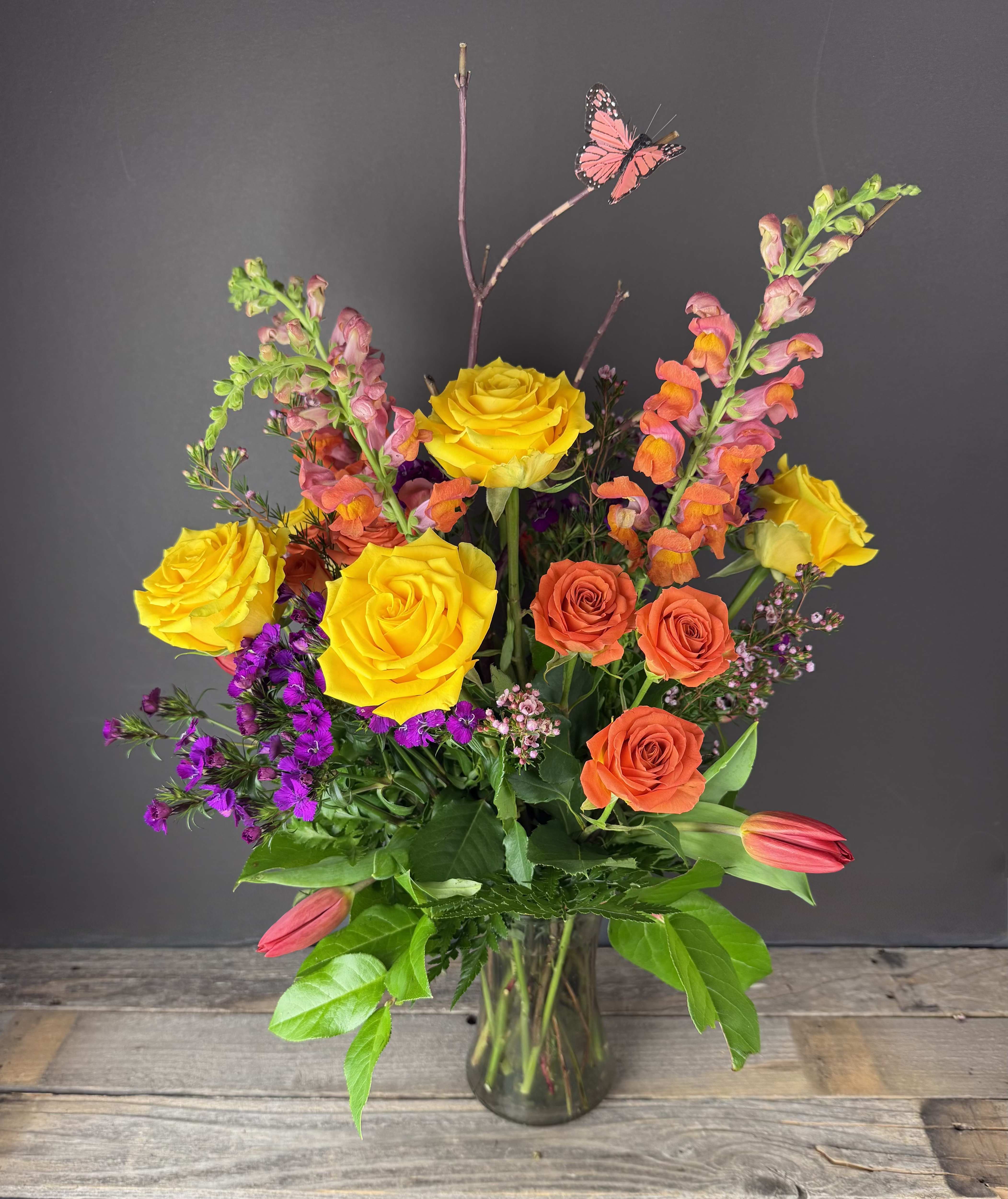 Tall mixed bouquet of yellow and orange roses with snapdragons, purple blooms and tulips in a clear glass vase