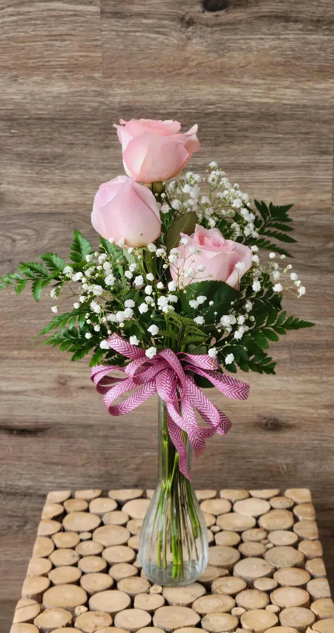 Pink Triple Rose Bud Vase - A clear glass bud vase filled with pink roses, baby’s breath, leather leaf fern, and a bow.