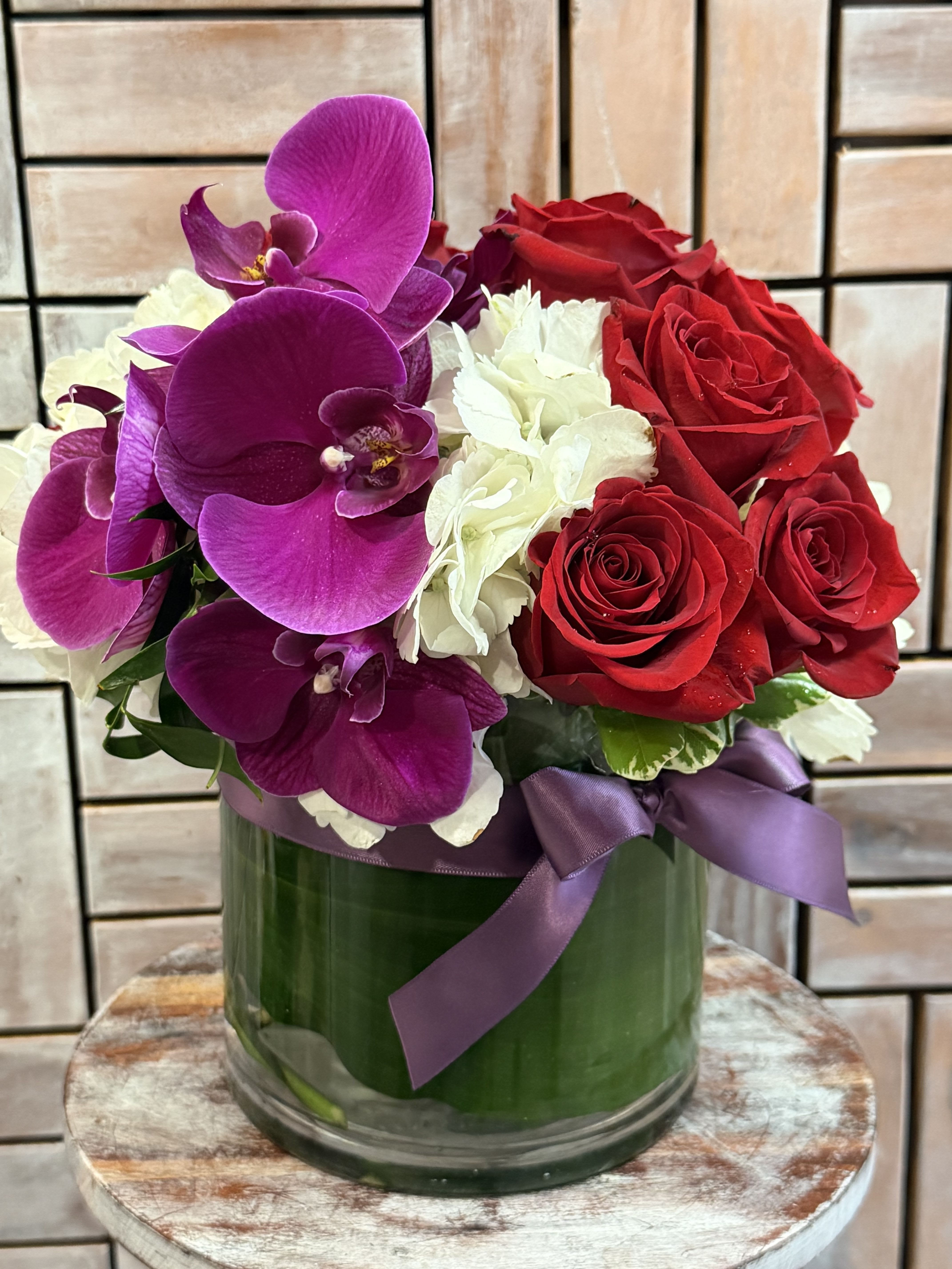 Velvet Orchid Romance - A luxurious arrangement featuring vibrant purple orchids, premium red roses, and soft white hydrangeas designed in a modern 6-inch glass cylinder with elegant leaf lining and satin ribbon detail. This romantic design blends bold color with timeless elegance, making it perfect for anniversaries, birthdays, Valentine’s Day, or any special occasion.
