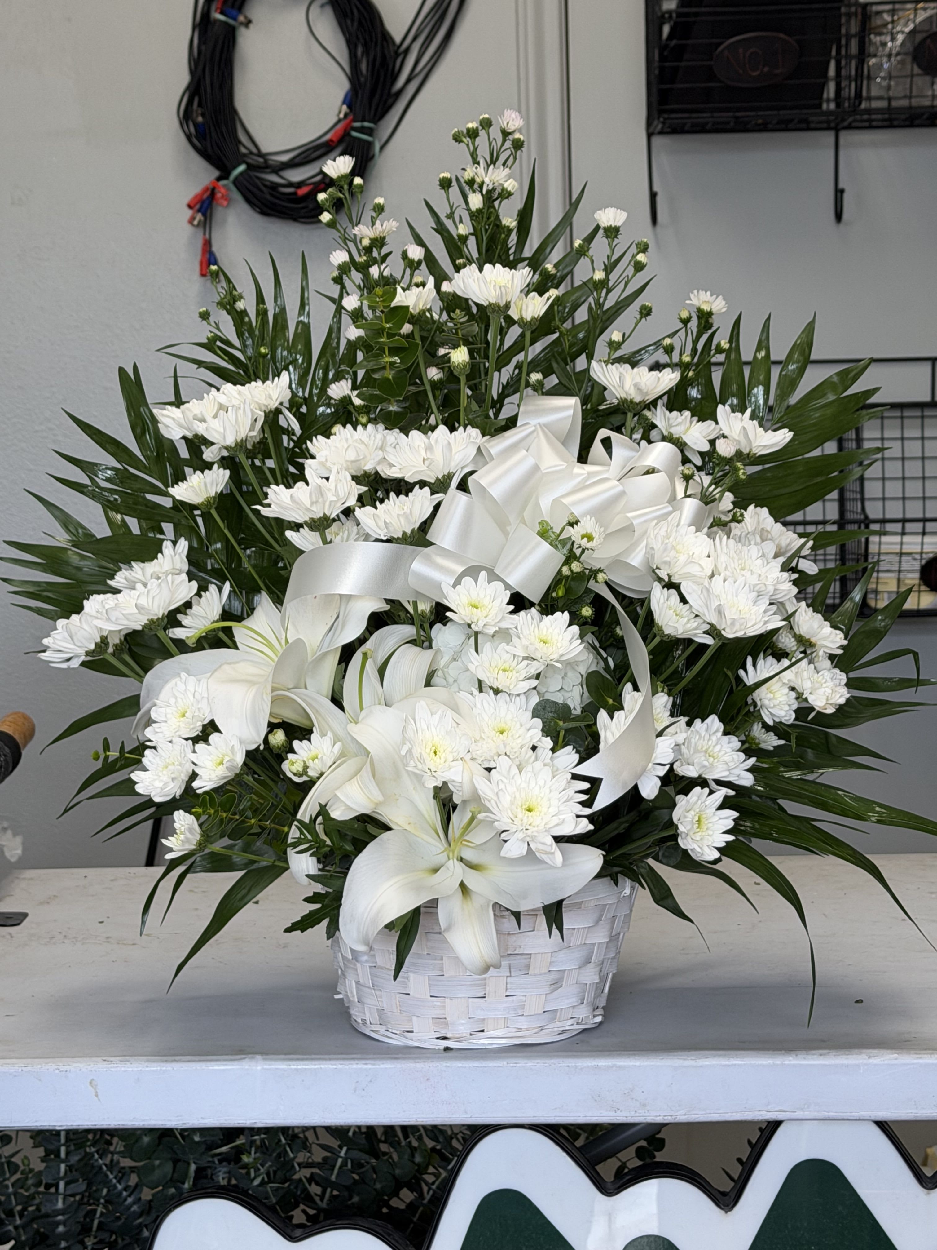 Cushion Sympathy basket - A calm cluster of white lilies and chrysanthemums arranged in a white woven basket makes this sympathy basket a serene way to offer comfort and remembrance. Designed as a memorial or sympathy arrangement, the all-white palette and wide white ribbon give it a formal, respectful presence that reads as quietly elegant for a funeral service or as a thoughtful condolence gift.  The cushion-style design is densely packed with large white lilies and clusters of white chrysanthemum blooms set against long, dark green palm fronds and complementary foliage that form a radiating, rounded silhouette. Finished with a crisp white ribbon and presented in a low white woven basket, the arrangement emphasizes texture and contrast-soft petals against pointed greenery-for a refined, peaceful look.  Ideal for honoring a loved one or sending condolences, this basket conveys sympathy and calm without being showy; it would also suit a formal occasion such as a wedding memorial or remembrance service. The compact, full shape sits comfortably on a memorial table or beside a casket; standard care is to keep flowers in a cool, sheltered spot and ensure the floral foam remains moist for longevity.