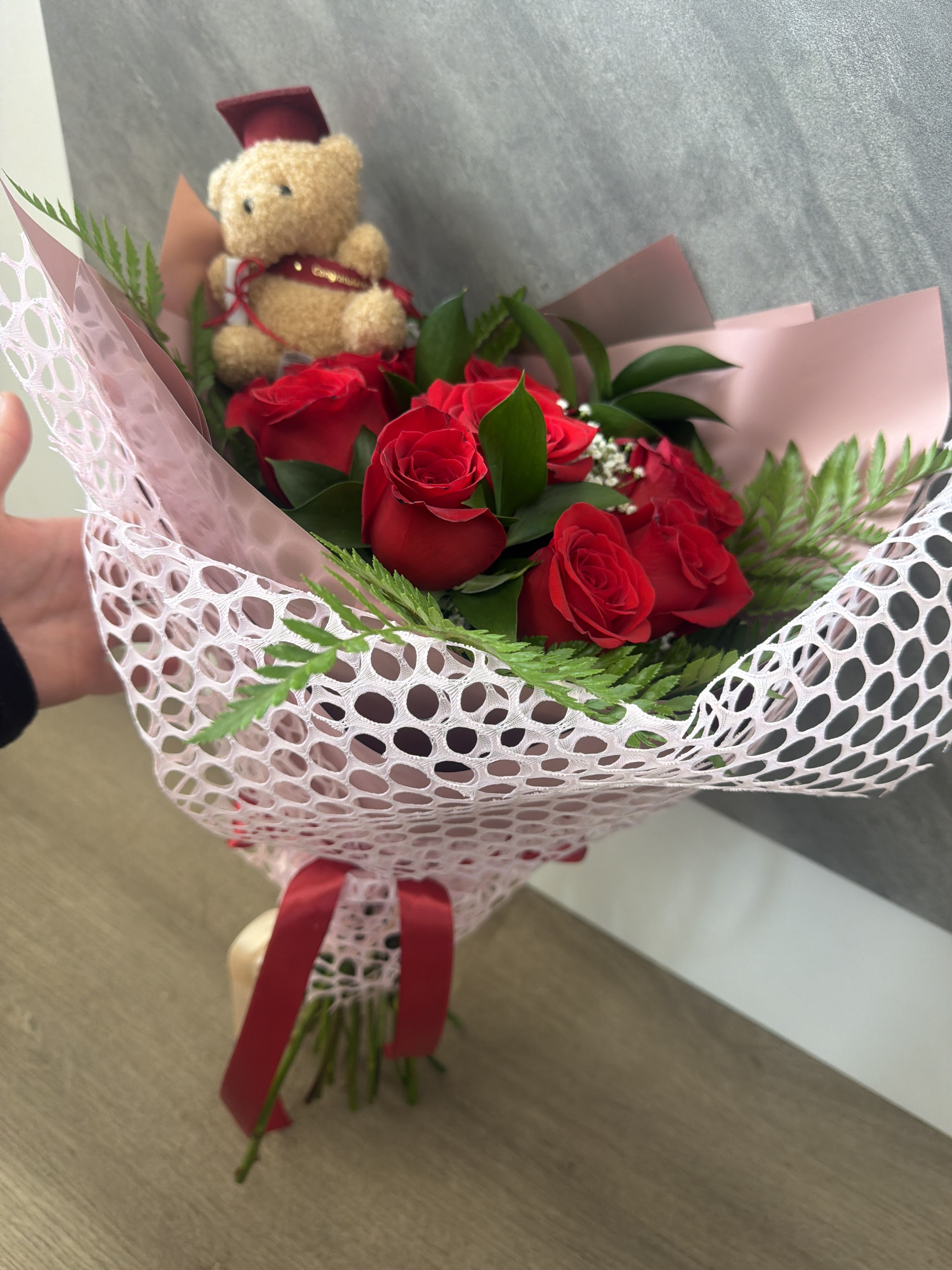 Graduation bouquet - Bright, tightly clustered red roses form the heart of this graduation bouquet, their velvety petals arranged around a small teddy bear wearing a graduation cap for a playful, congratulatory touch. Designed as a graduation gift that also reads romantic and celebratory, this arrangement works beautifully for graduation, birthday, anniversary, or "congrats" moments-she'll feel seen and celebrated the instant she unwraps it.  The bouquet is wrapped in pale pink paper with a white mesh overlay and finished with a red ribbon, creating a polished, gift-ready presentation. Choose the clear glass vase option for an added keepsake look-the vase is tied with the same red ribbon and elevates the arrangement for display. Colors in the design include rich red, soft pink, white accents and greenery for contrast; materials include the plush teddy bear, decorative wrapping and ribbon.  Perfect for graduates, a sweetheart, or anyone you want to honor on their big day, this bouquet ships ready to present and makes a memorable table centerpiece or dorm-room surprise. If you'd like vase delivery, select that option at checkout; to keep blooms fresh, change the water every 2-3 days and trim stems slightly when you refresh the water.